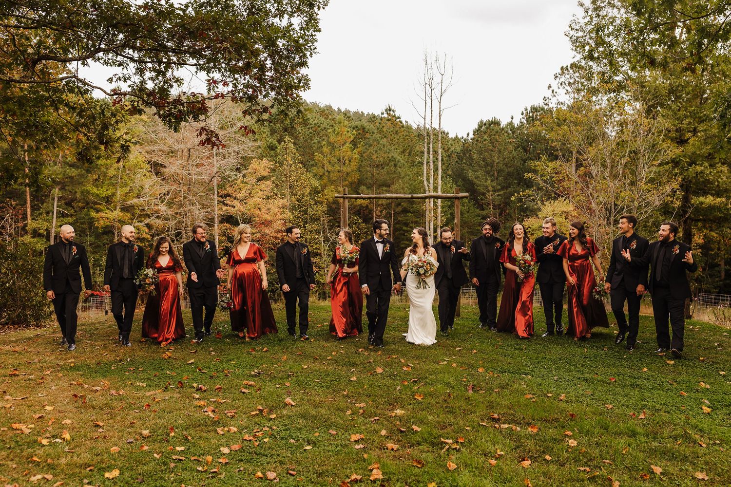 Wedding party in burgundy and black attire stands on autumn lawn with colorful fall foliage in background.