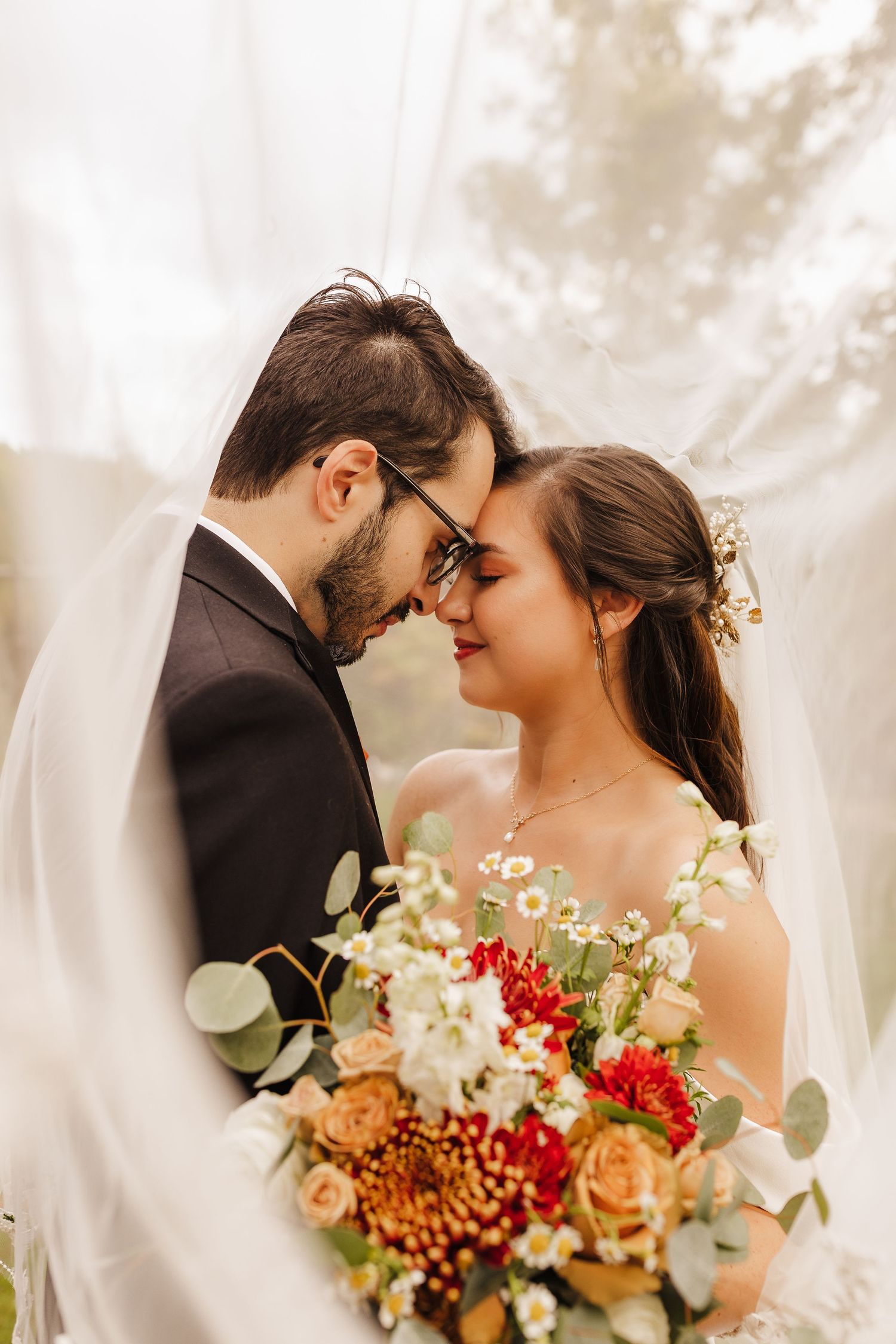 Romantic wedding couple embraces with autumn-inspired bouquet of red and cream flowers against soft white backdrop.