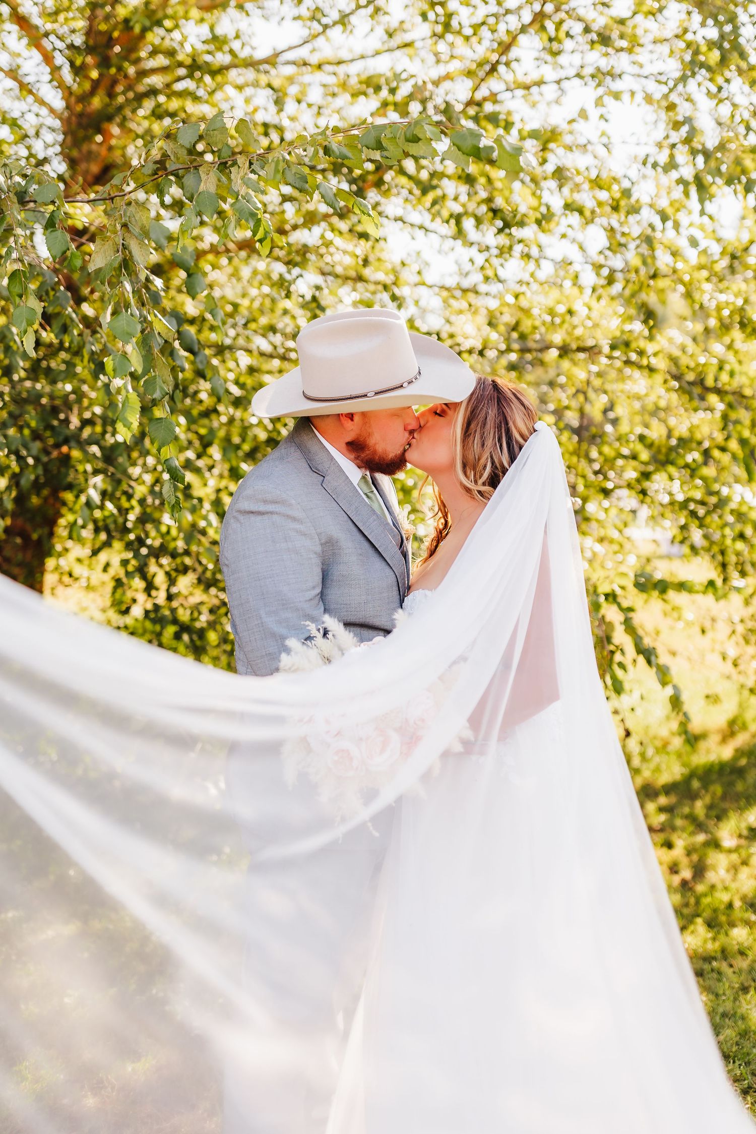 Bride and groom share intimate moment under trees with flowing white veil in natural setting.