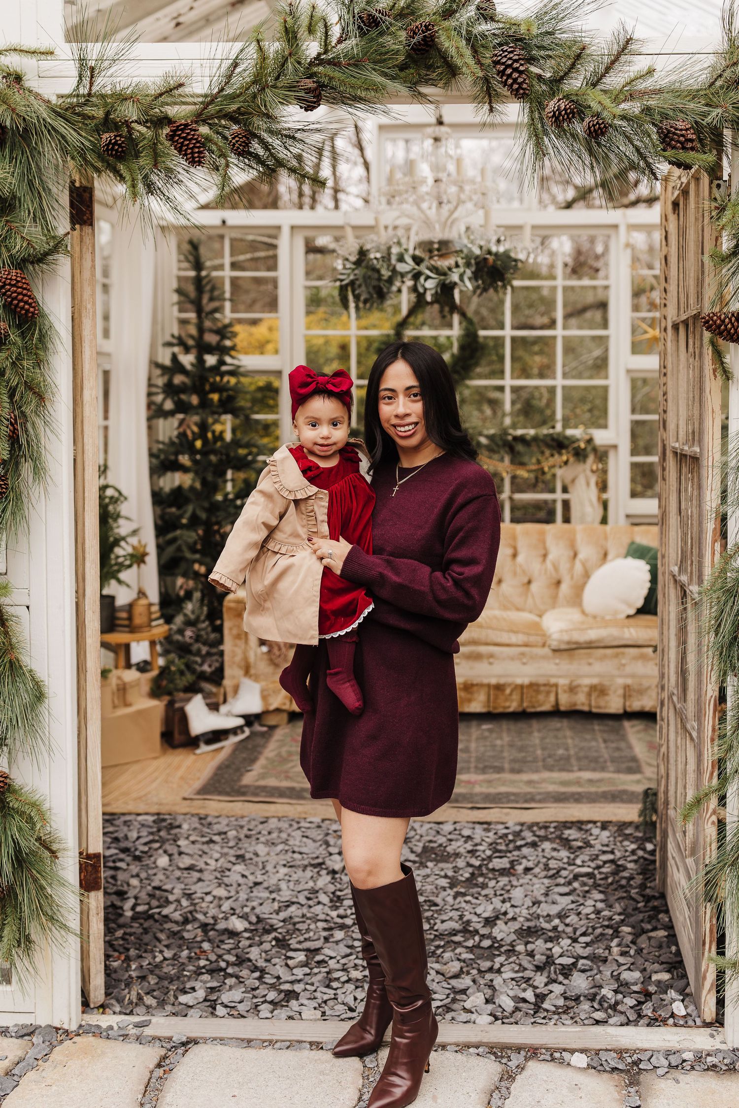 Woman in burgundy dress holding toddler girl in red bow, posing in Christmas greenhouse doorway with pine garland.