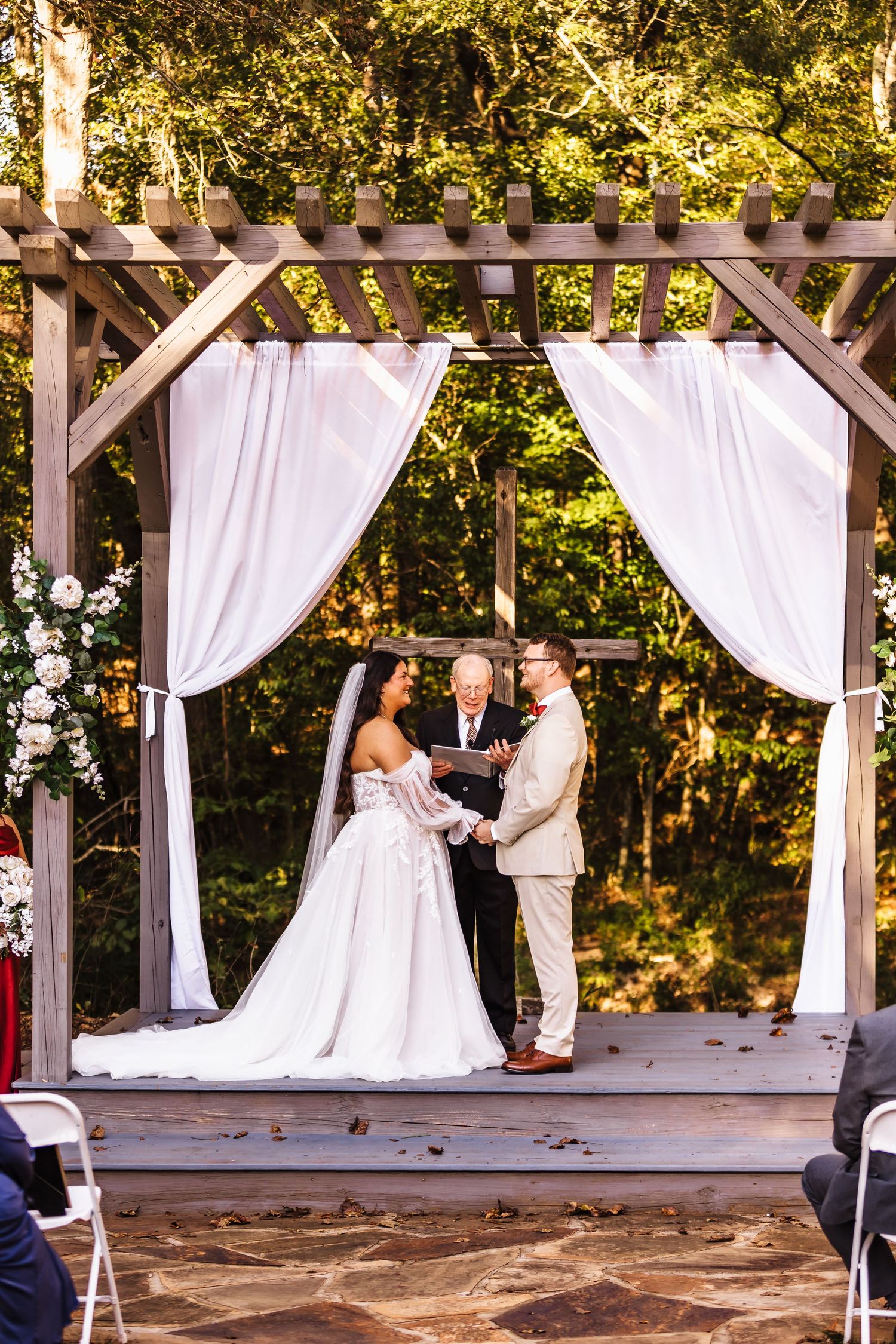 Bride and groom exchange vows under a wooden pergola with white draping, surrounded by lush greenery at an outdoor wedding.