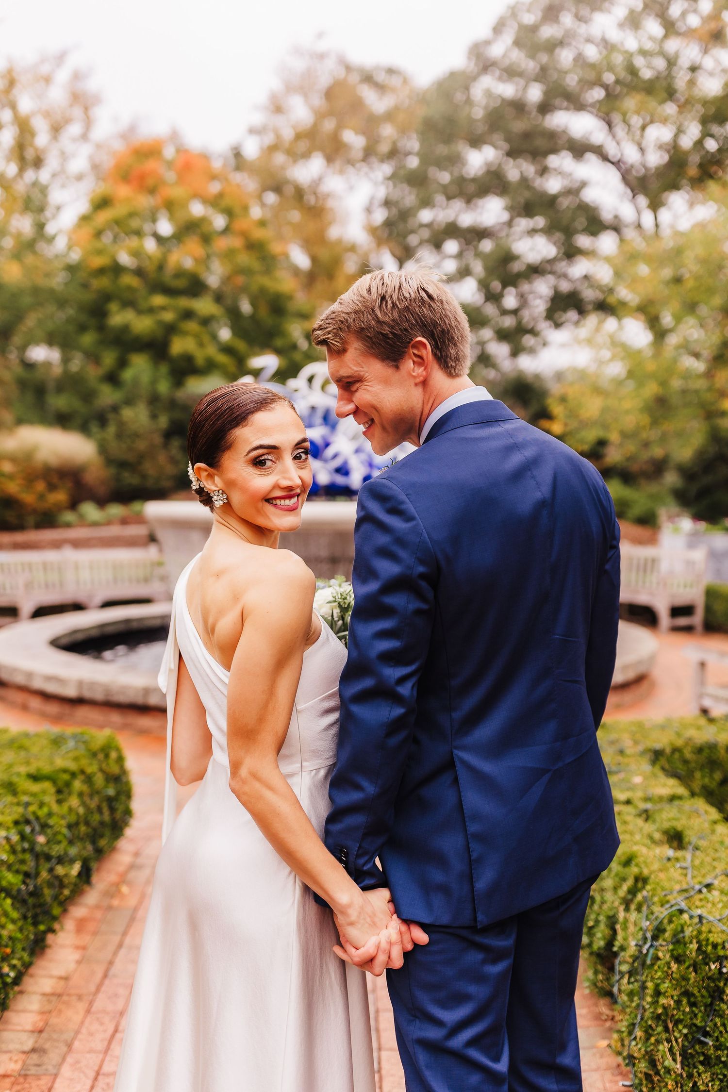 A romantic wedding portrait shows a couple in formal attire standing on a garden path during autumn.