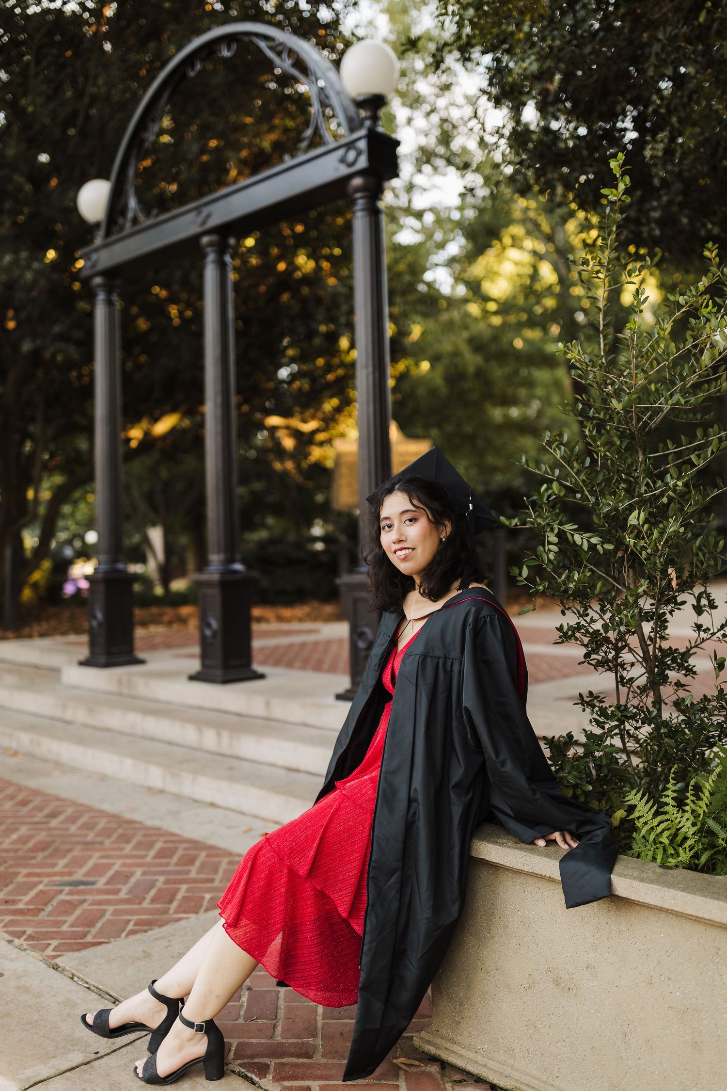 Graduate in cap and gown sits on ledge by iconic University of Georgia arch during sunset graduation photoshoot.