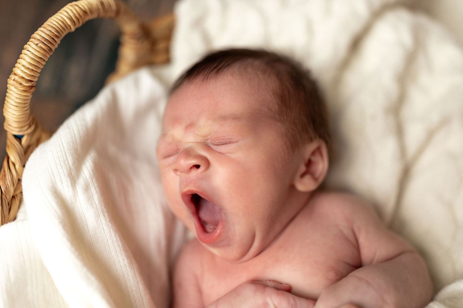 A newborn baby yawns while lying on white bedding in natural lighting.