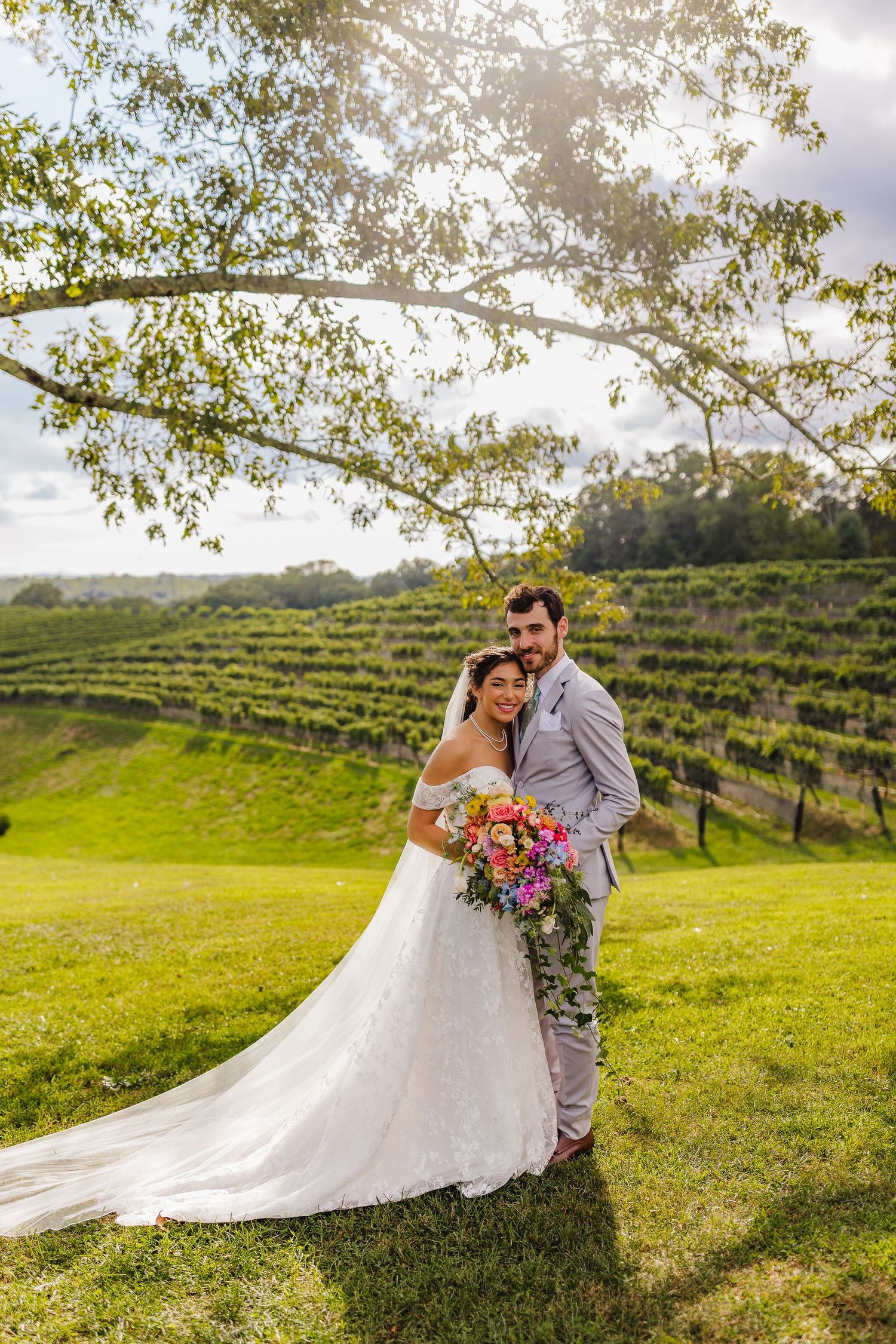 Bride and groom pose in front of lush vineyard rows under a tree, bride holding a vibrant cascading bouquet.