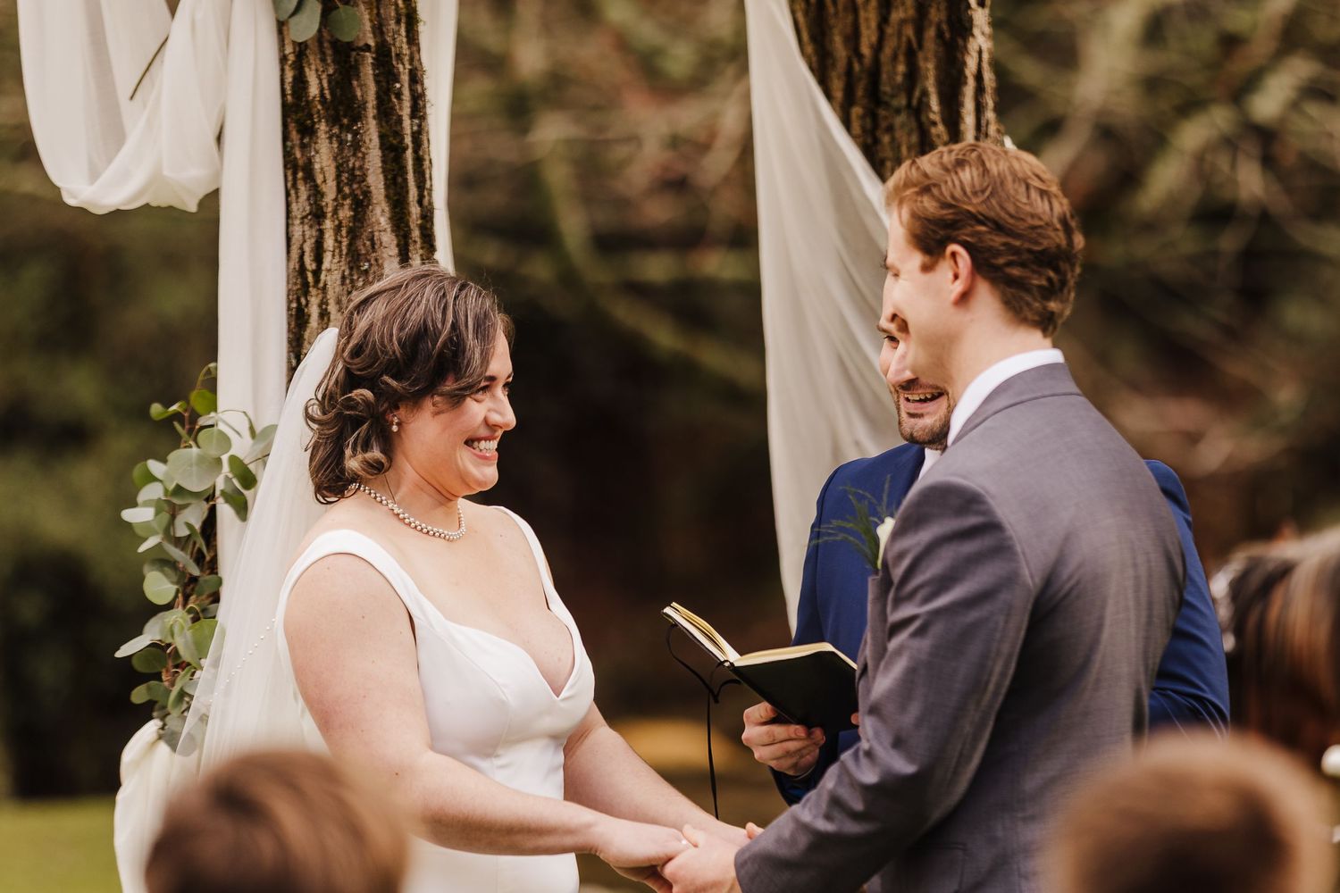 Bride and groom smiling at each other during outdoor wedding ceremony, exchanging vows under draped floral arch.