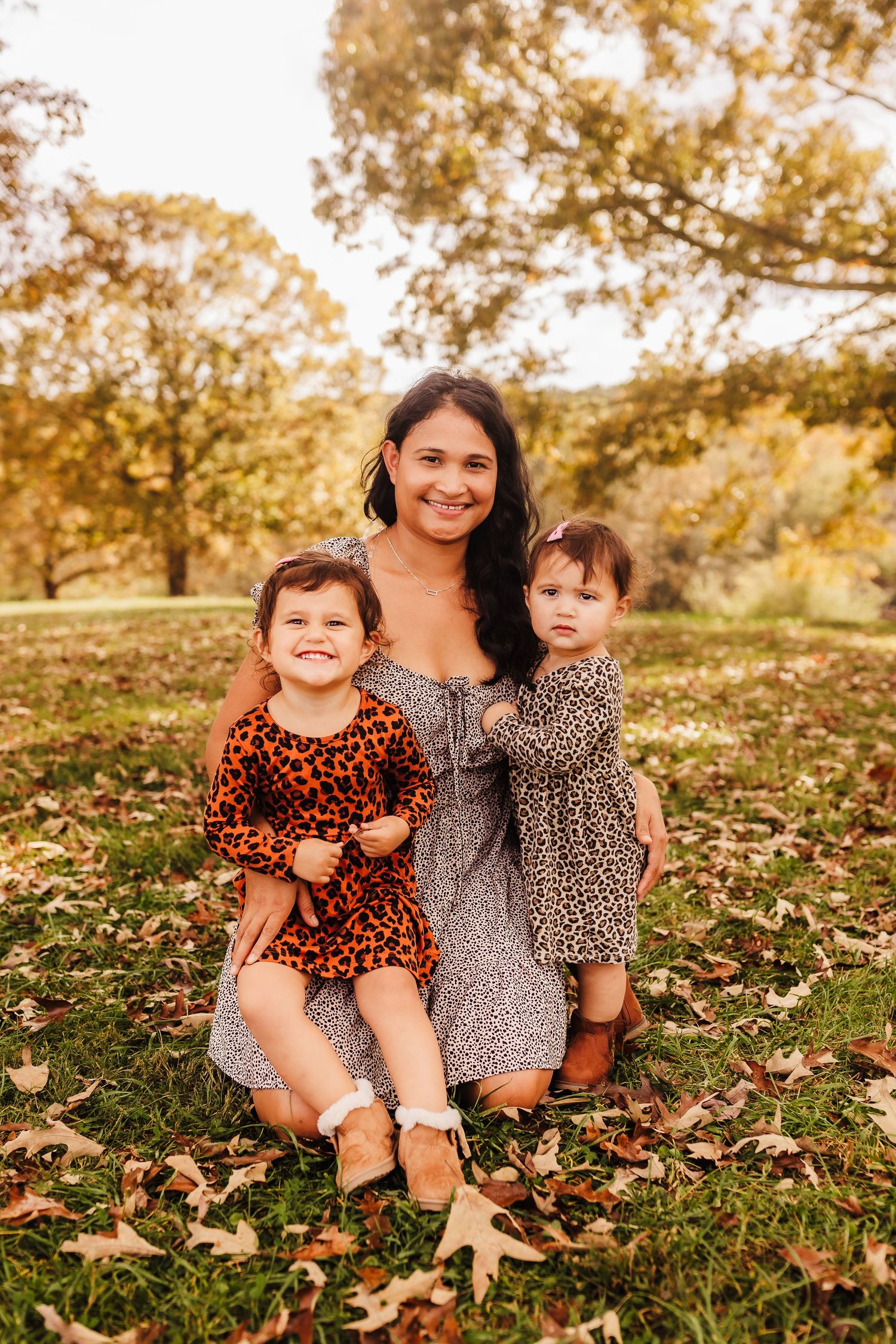 Mother in floral dress sitting with two young children in matching outfits among autumn leaves.
