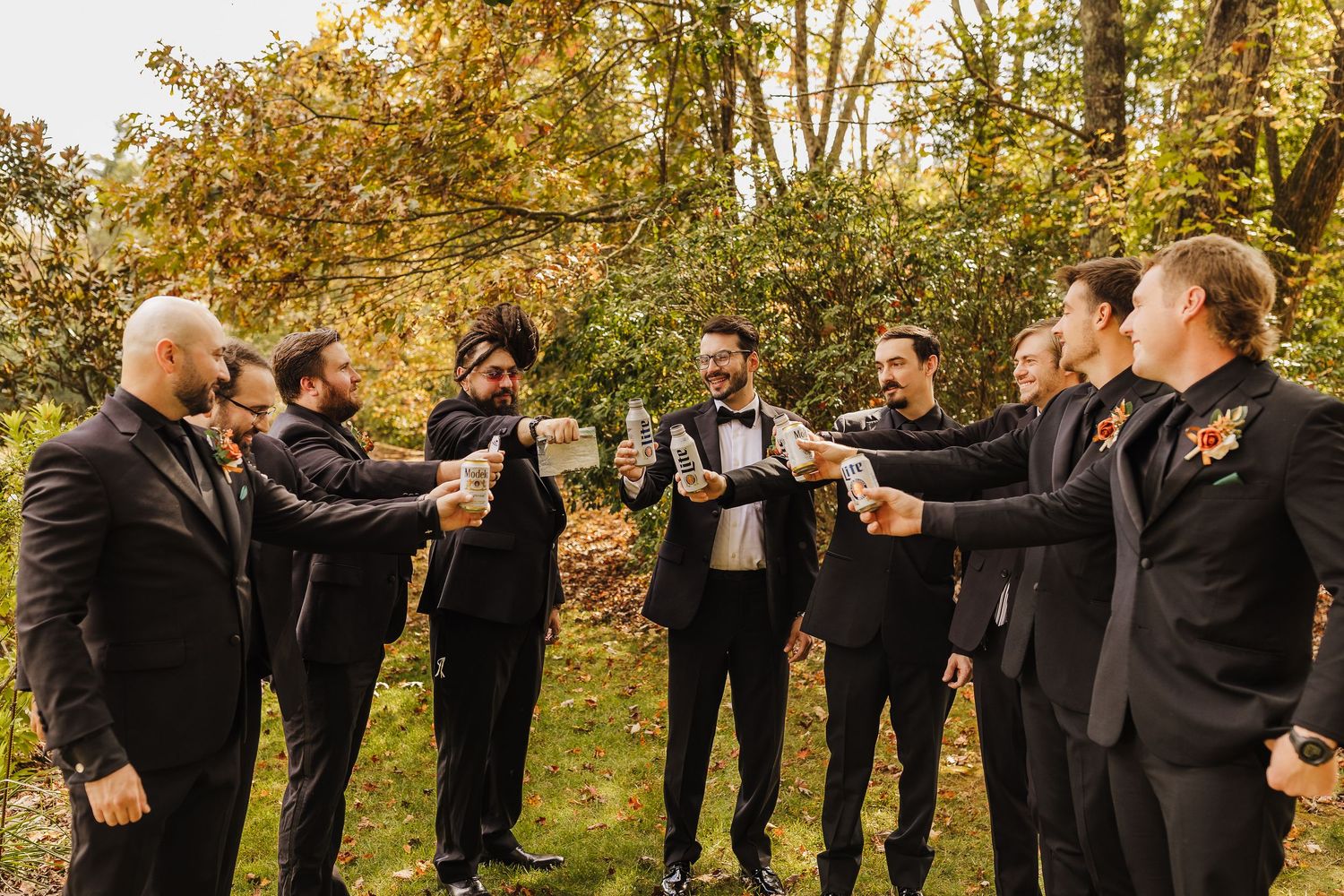 Groomsmen in black suits pose together in a line against a backdrop of golden fall trees.