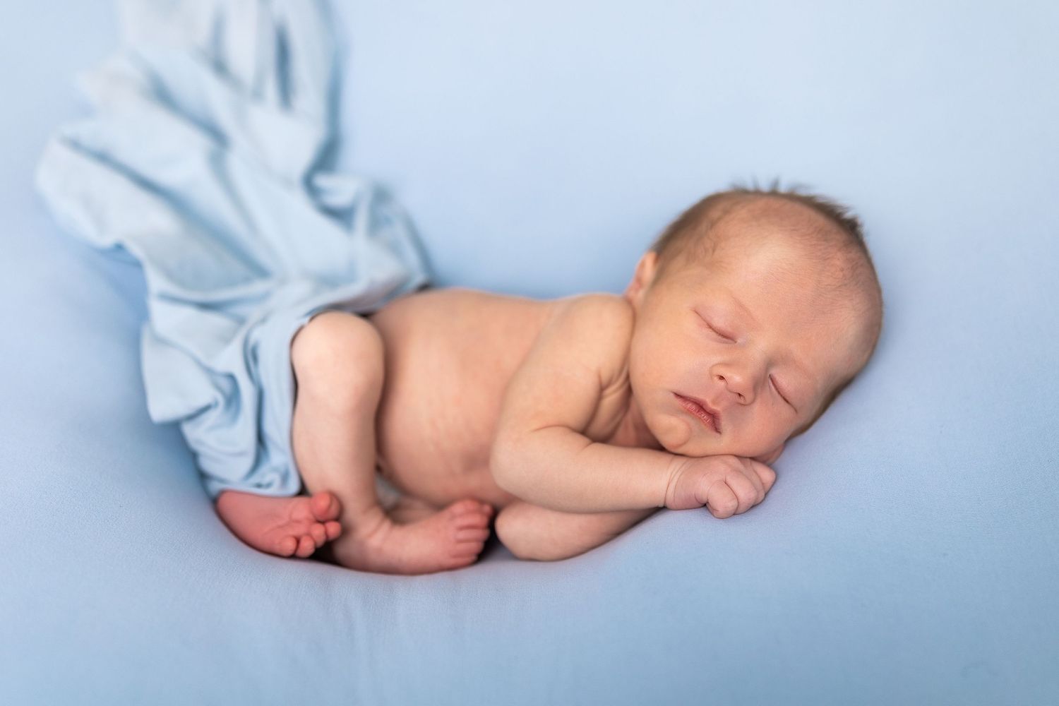A peaceful sleeping newborn baby wearing white lies on a soft blue blanket in a curled up position.