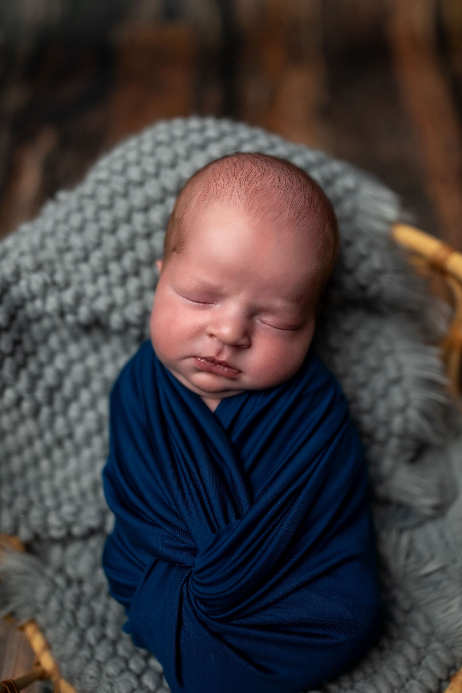 Peaceful sleeping newborn wrapped in navy blue swaddle blanket resting on soft gray knitted blanket.