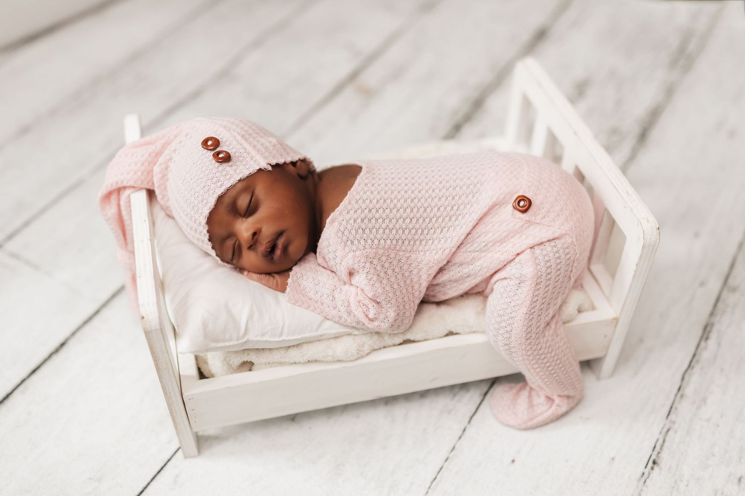 A newborn baby wearing pink sleepwear lies peacefully on white wooden flooring during a photography session.