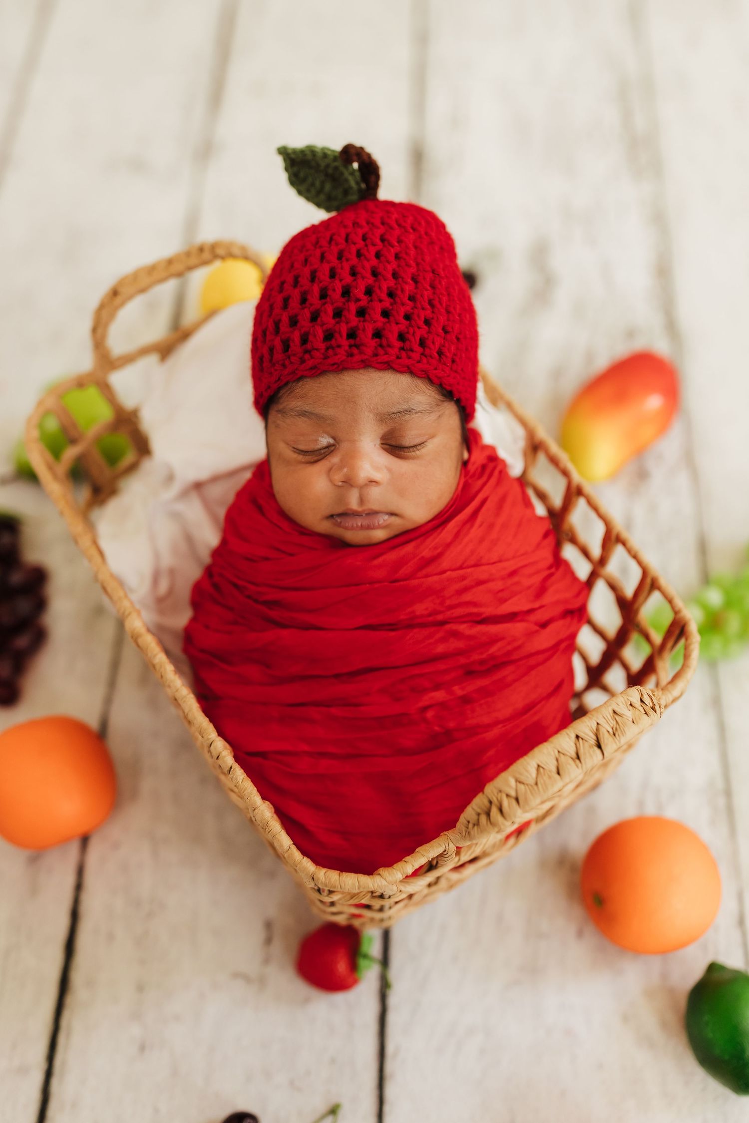 A sleeping baby wrapped in red knit fabric rests in a woven basket beside colorful citrus fruits on a white wooden surface.