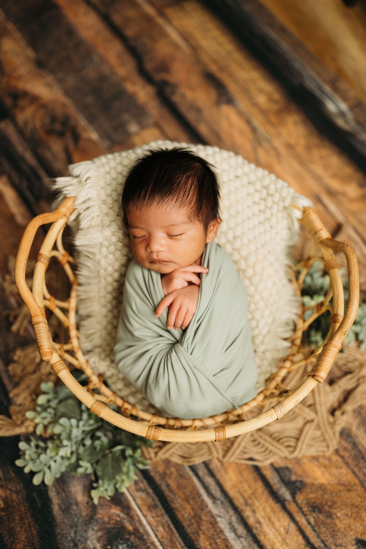 Sleeping newborn baby wrapped in sage green swaddle rests peacefully in round wicker basket with eucalyptus decor.