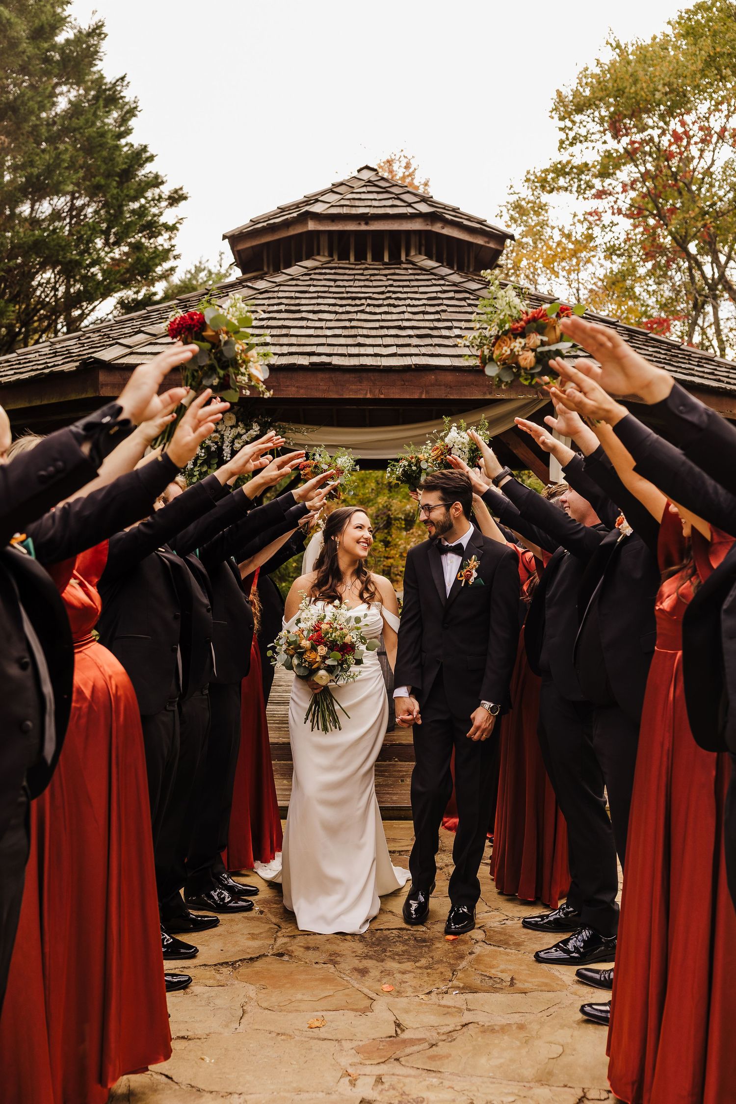 Wedding party forms a tunnel with raised arms as newlyweds walk through in front of rustic building.