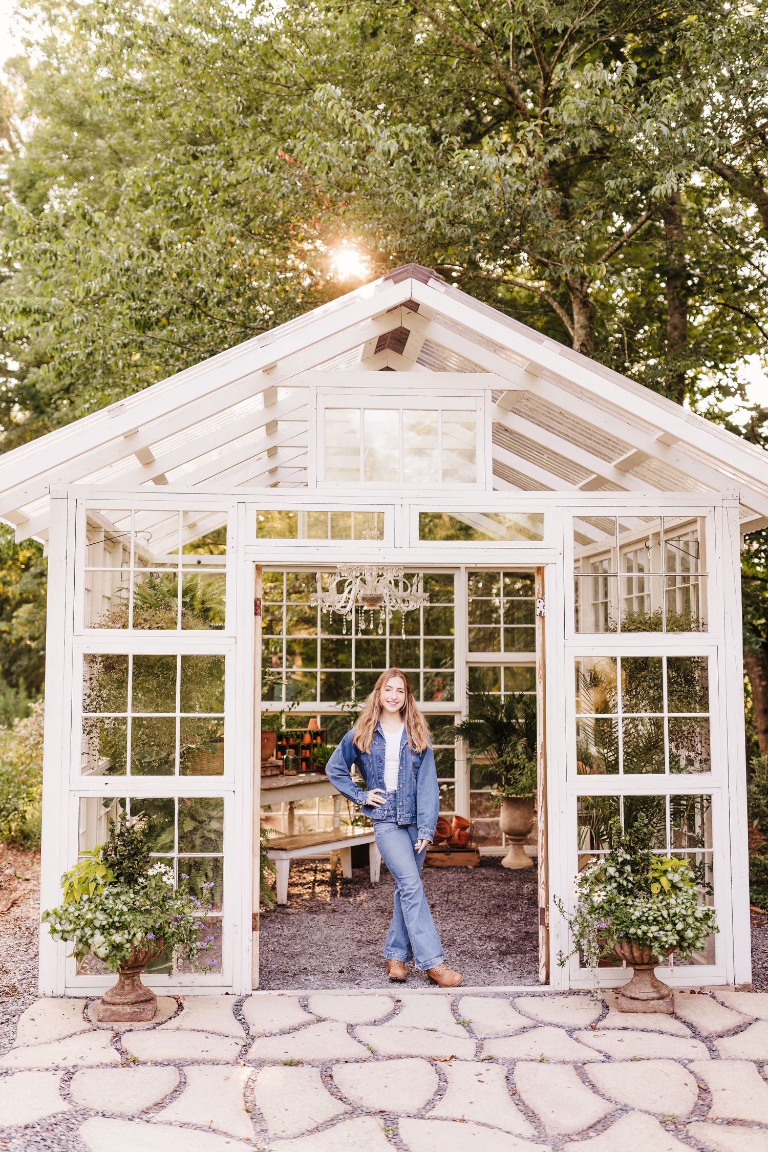 Woman in denim outfit standing in doorway of charming white greenhouse with large windows surrounded by lush greenery.