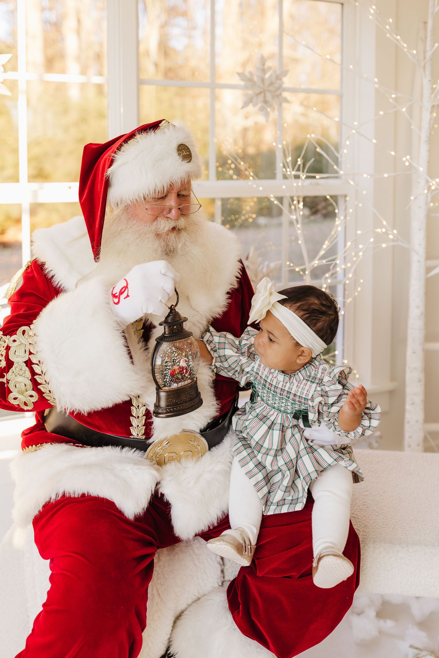 Baby in plaid outfit sits on Santa's lap, examining a snow globe lantern, with festive white lights in background.