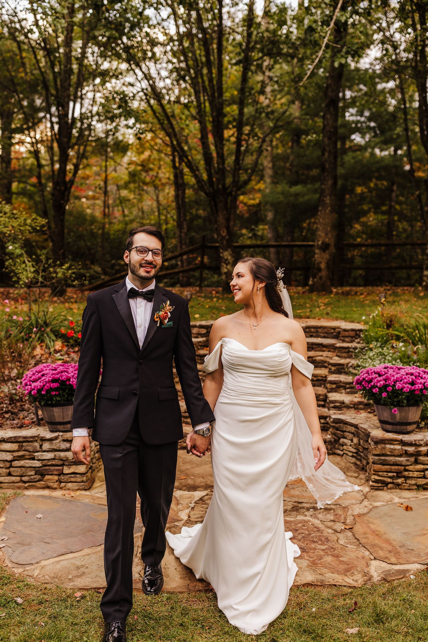 Newlyweds walking hand in hand along a stone path with pink flowers and autumn trees in background.
