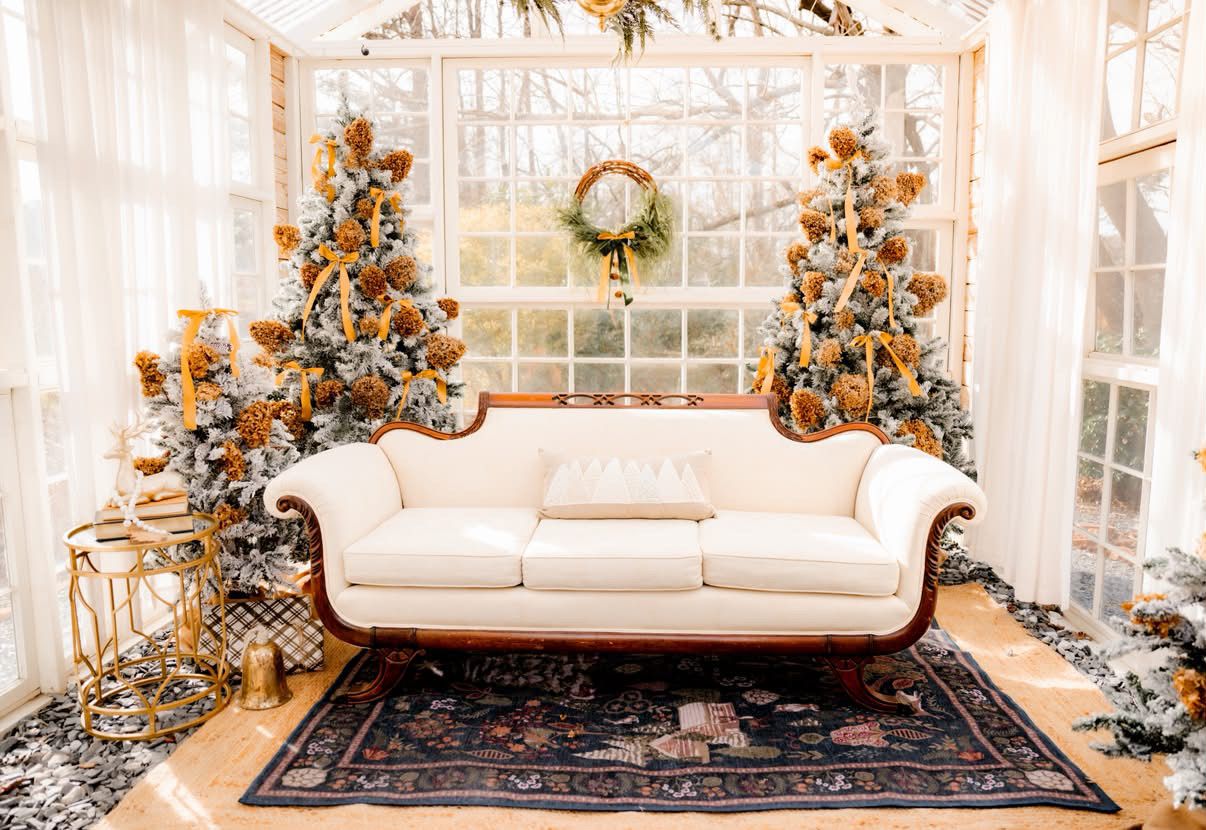 Elegant white sofa flanked by gold-decorated Christmas trees on a Persian rug in a bright sunroom.