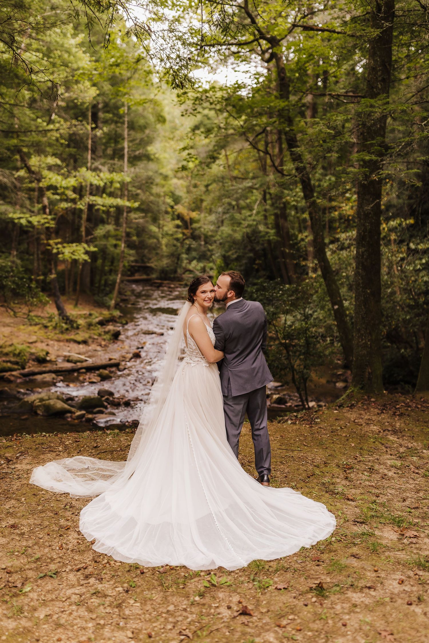 Bride and groom embrace near a forest stream, her long white gown trailing elegantly on the ground.