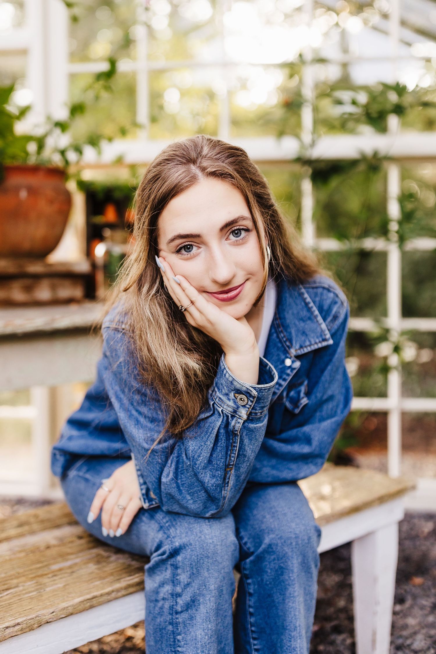 Young woman in denim jacket sitting on bench, smiling with hand on cheek, greenhouse background.