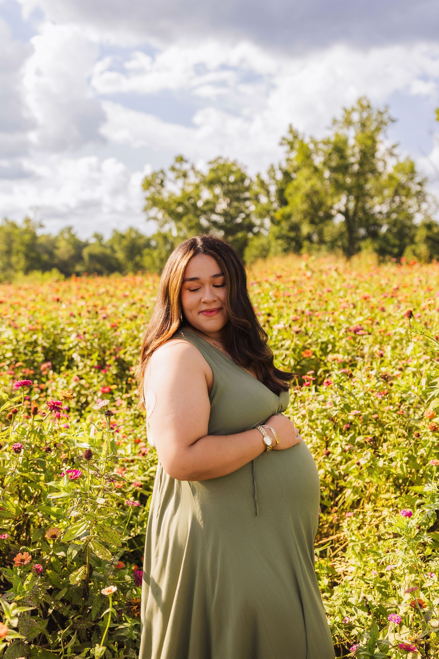 Pregnant woman in green dress cradling baby bump in vibrant flower field under cloudy sky.