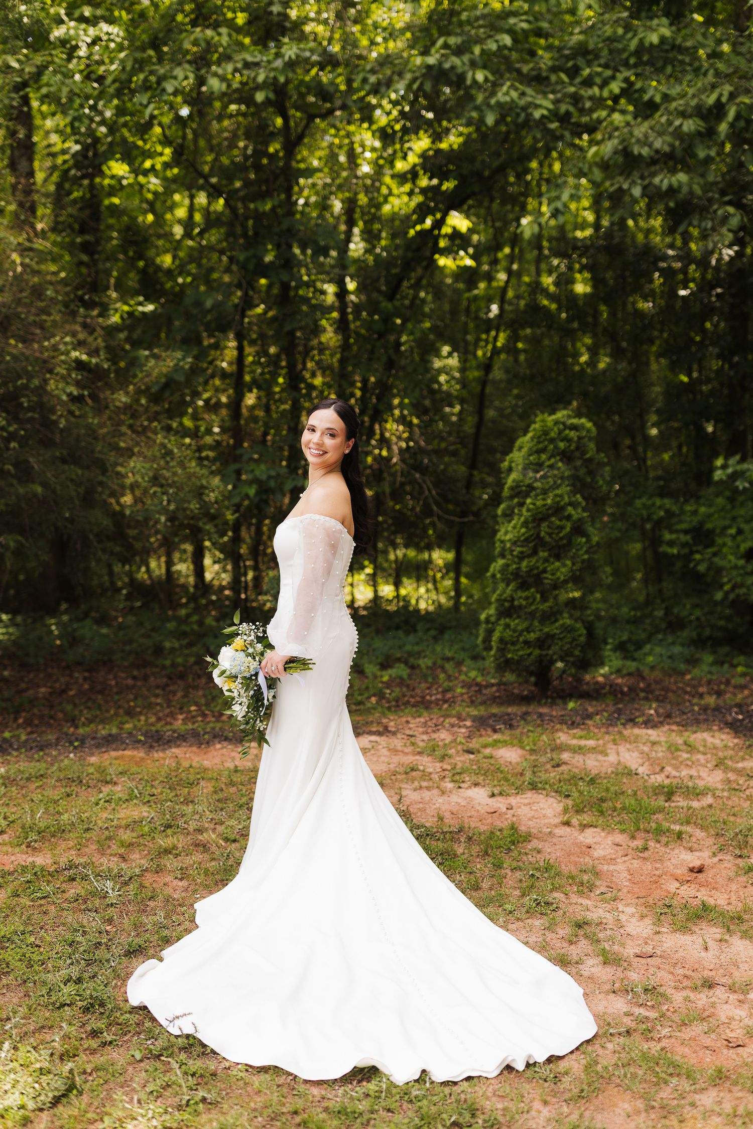 Bride in white mermaid gown with long train holds bouquet outdoors in lush green forest setting.