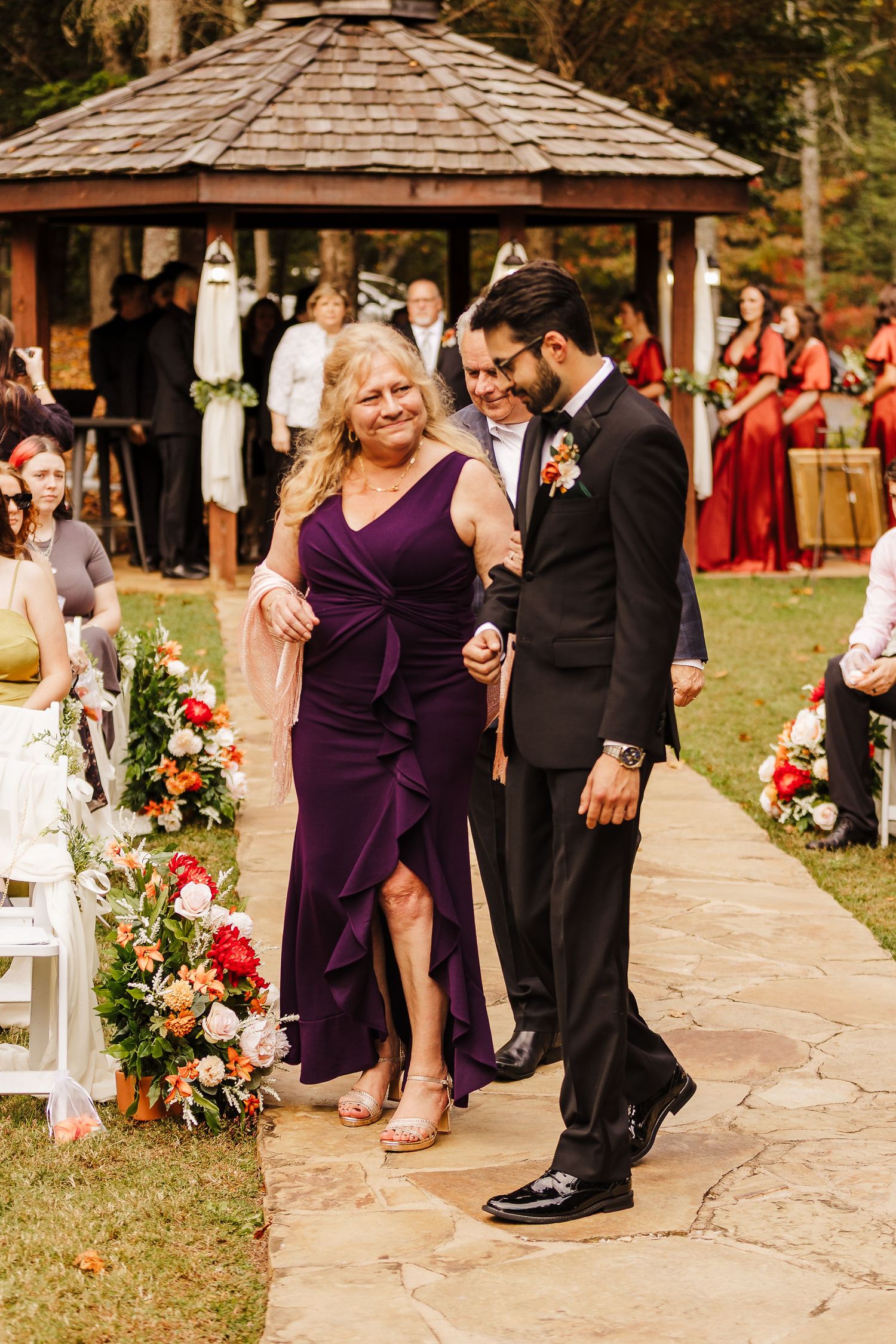 Wedding guests in purple dresses and formal attire gather along a stone pathway at an outdoor venue.