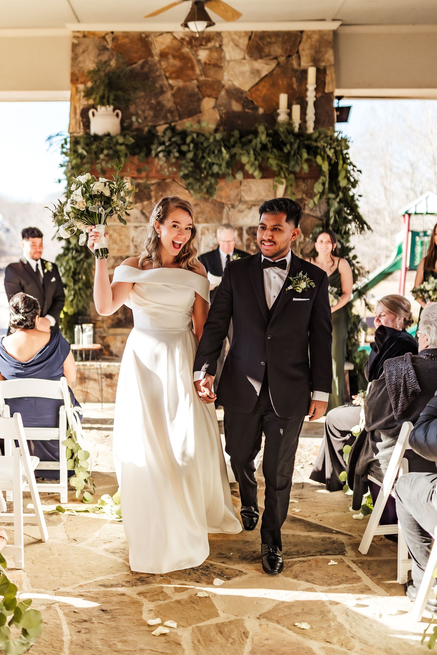 Joyful wedding recessional with decorated stone fireplace backdrop and white chairs.