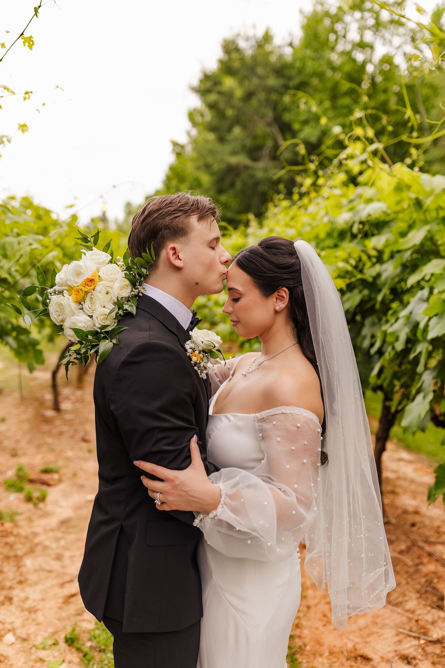Bride and groom share a romantic moment in a vineyard, with bride in white gown and veil holding a bouquet.