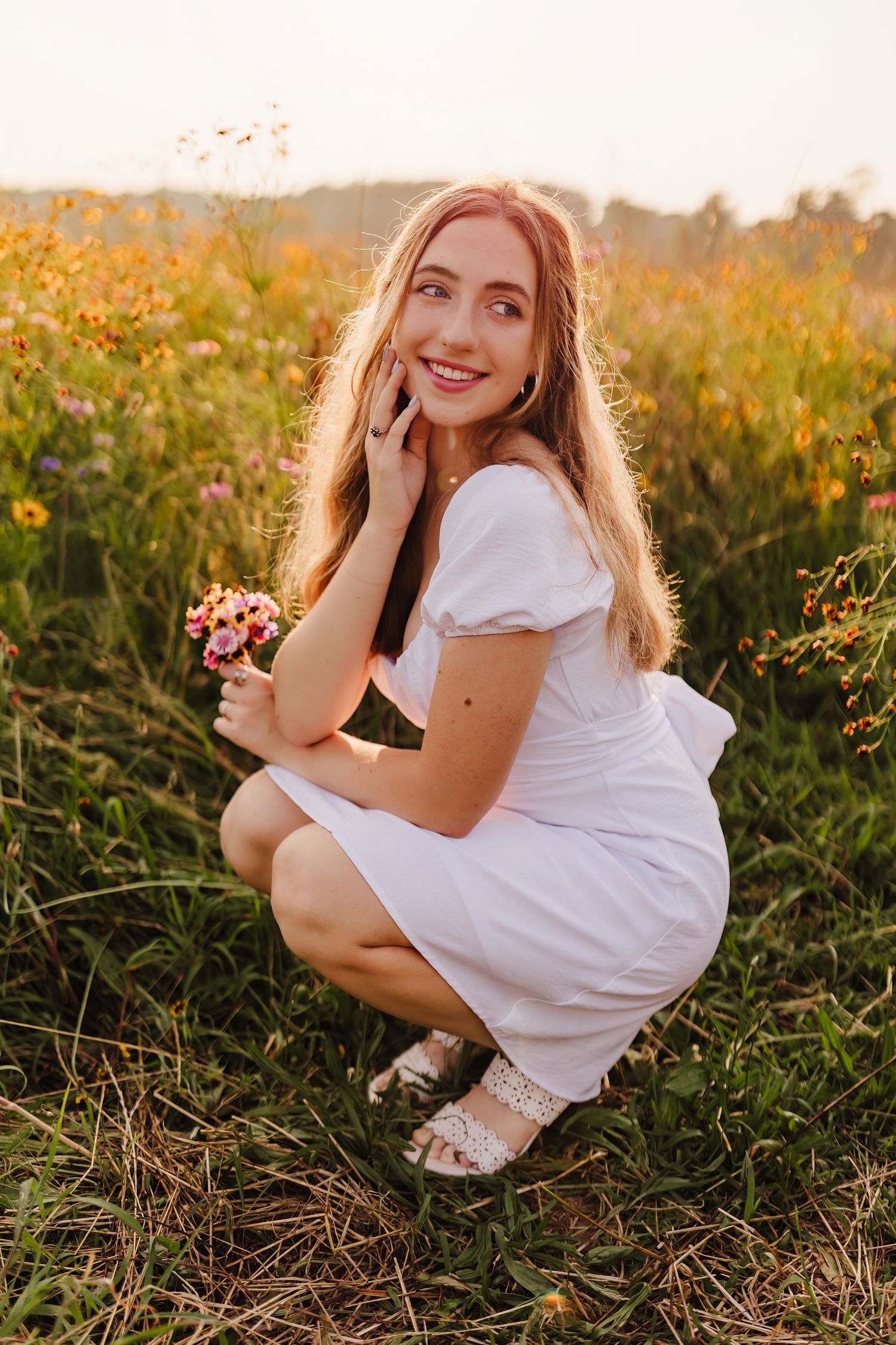 Smiling young woman in white dress crouching in a golden wildflower field holding small pink flowers.