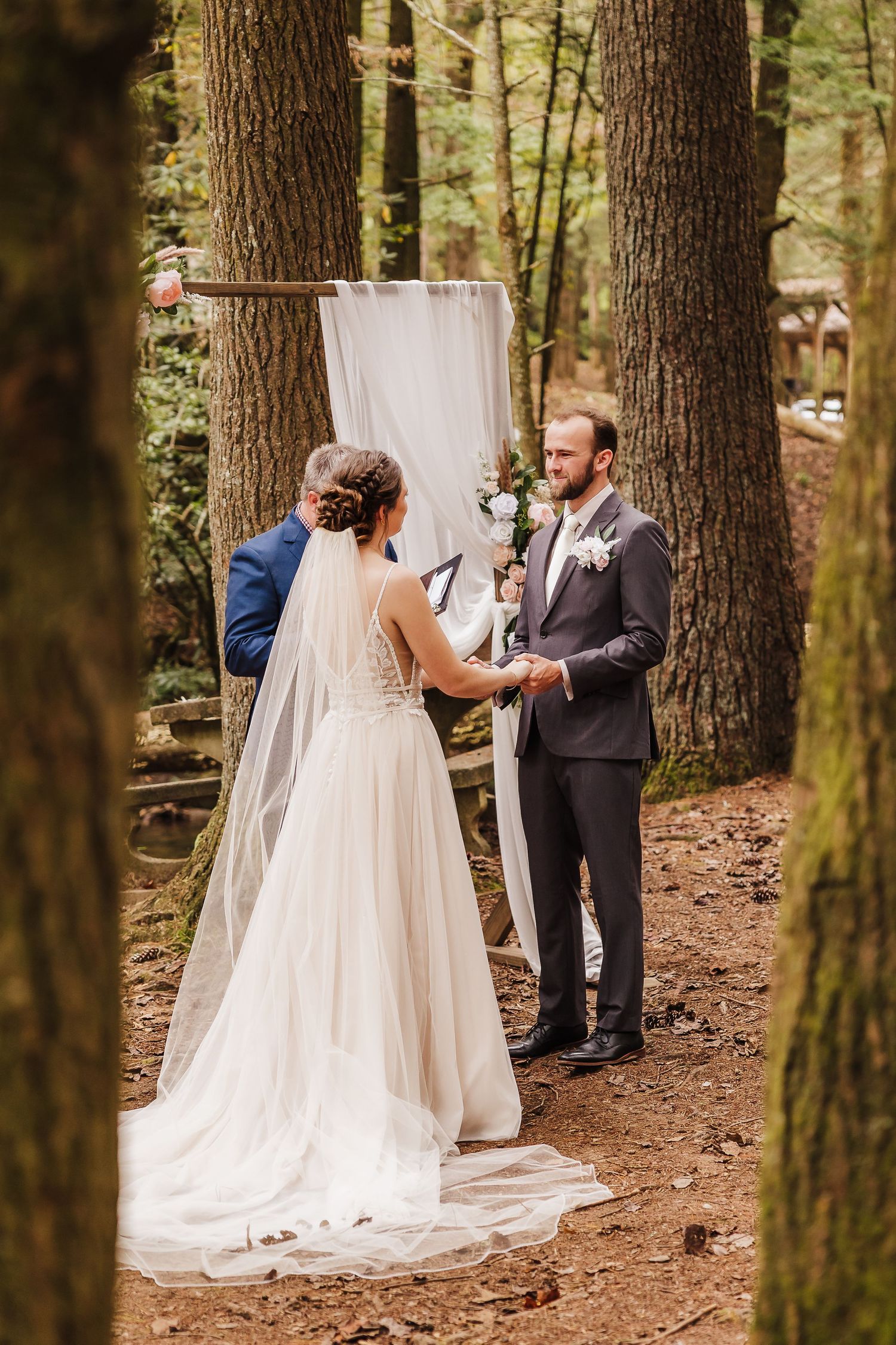Bride and groom exchange vows in a romantic outdoor forest wedding ceremony with a rustic floral arch backdrop.