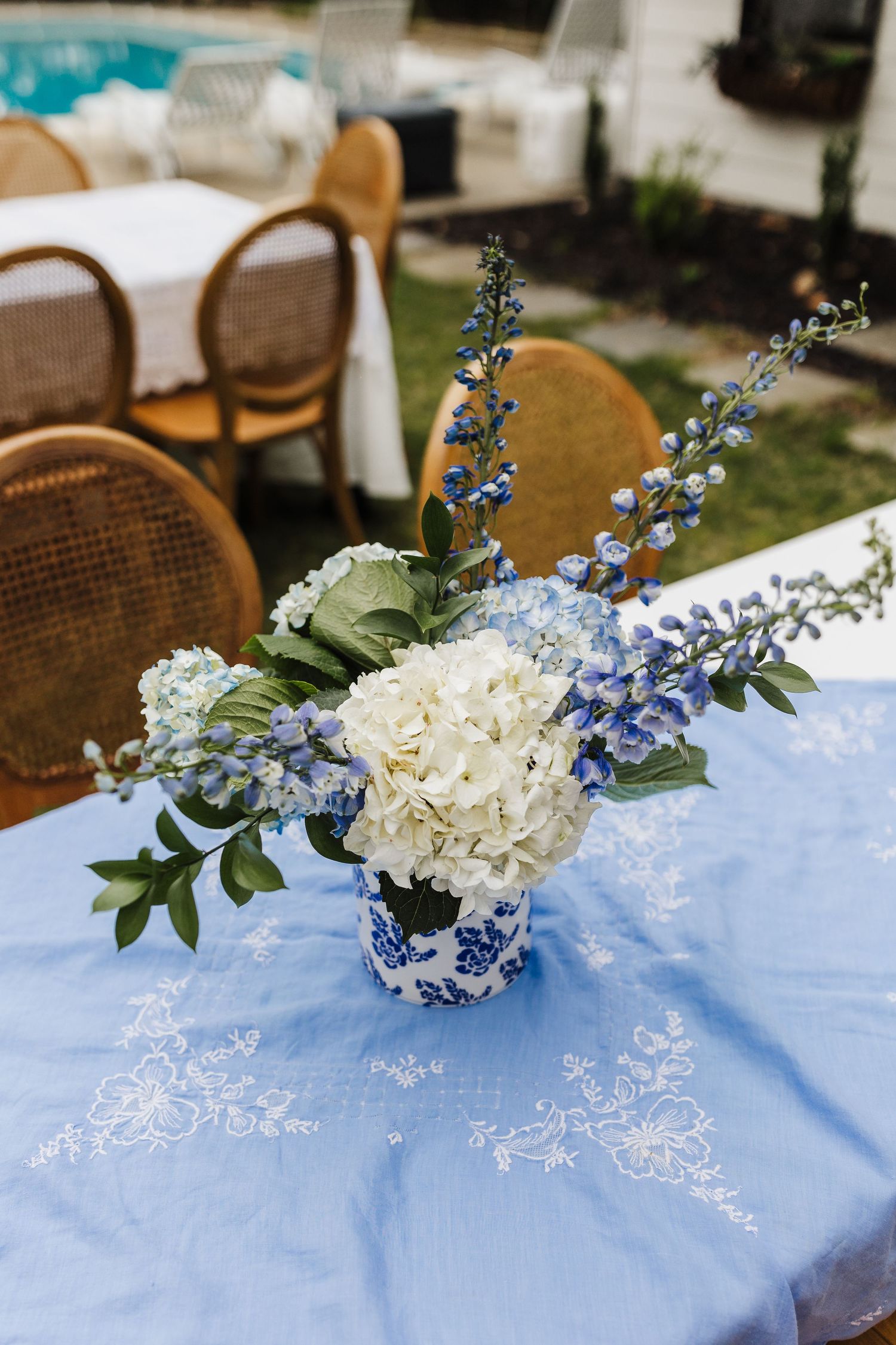 White hydrangeas and blue delphinium flowers arranged on a light blue tablecloth with delicate embroidered details.