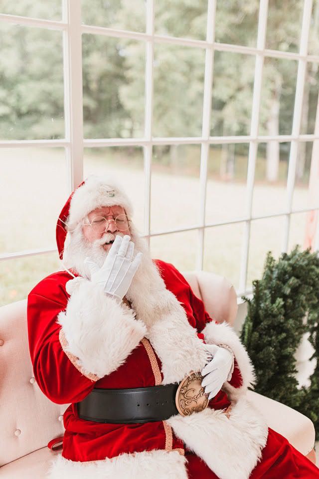 Santa Claus in red suit sitting in chair by large window, covering mouth with white gloved hand.