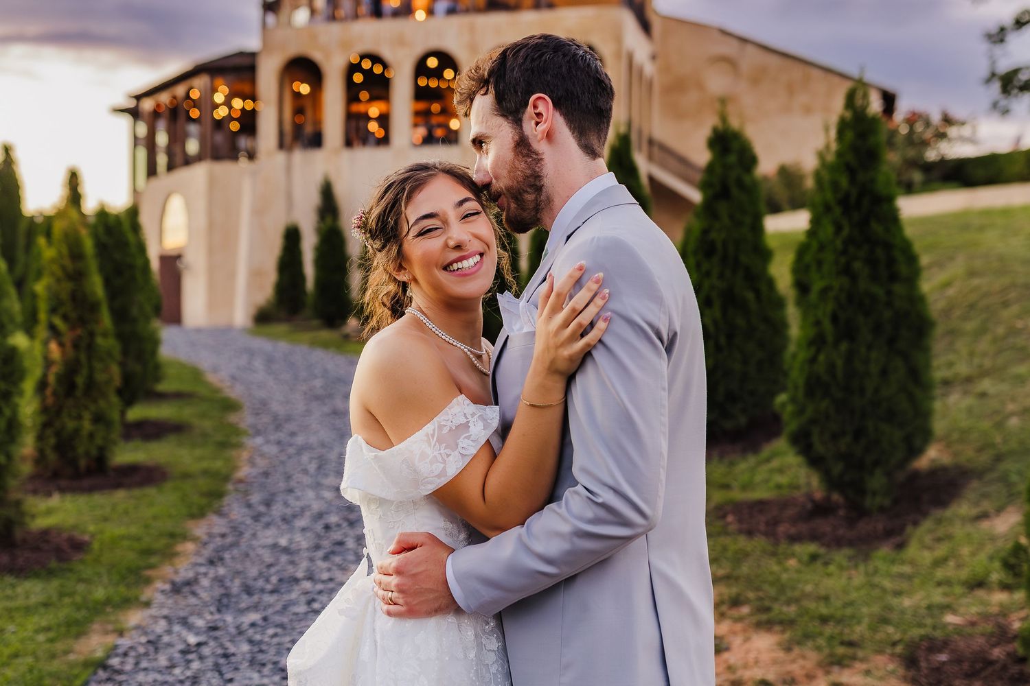 Bride and groom embrace outdoors at dusk near elegant venue with cypress trees at golden hour wedding.