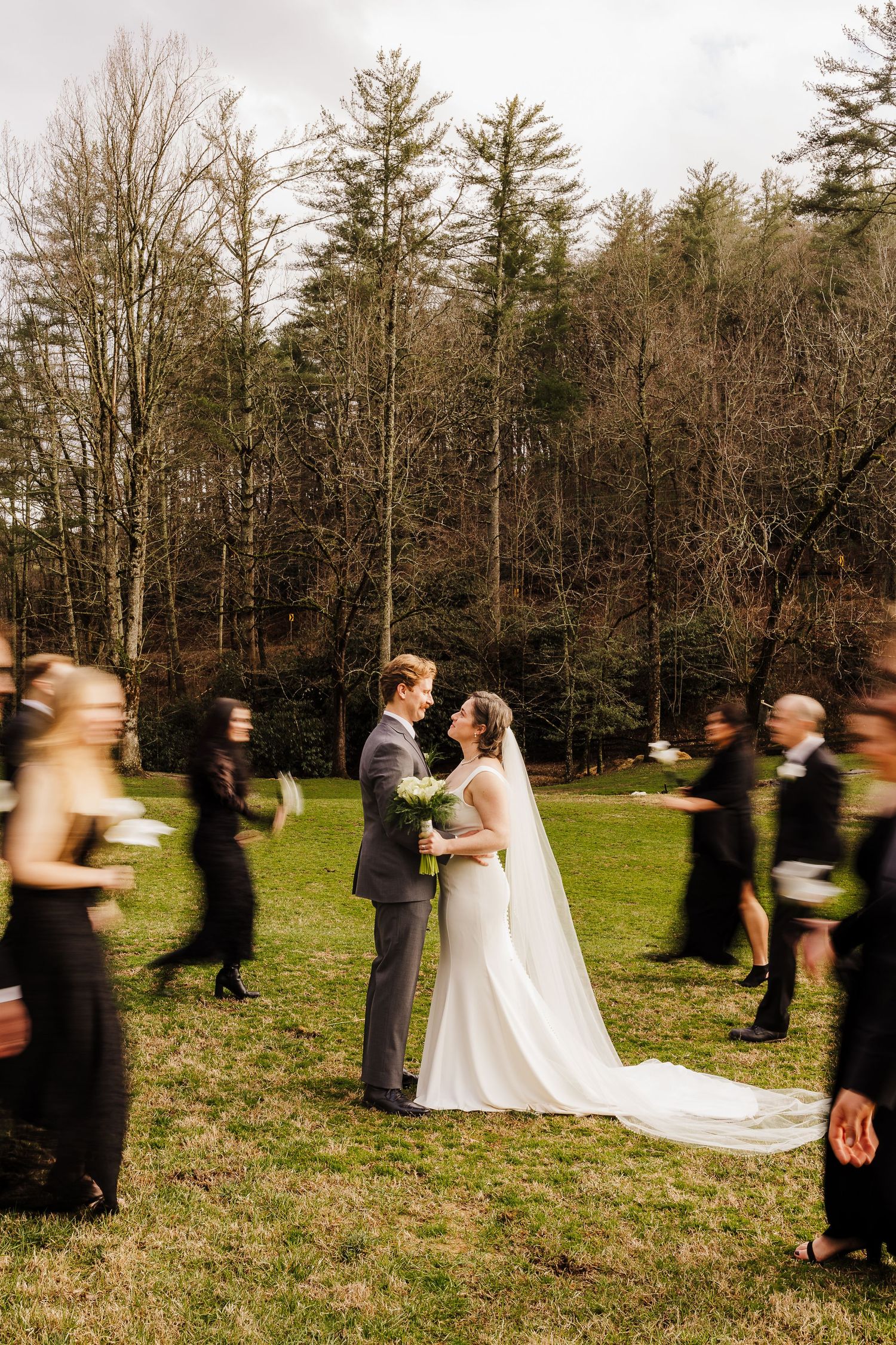 Newlyweds share romantic moment on green lawn as wedding party moves around them in motion blur.