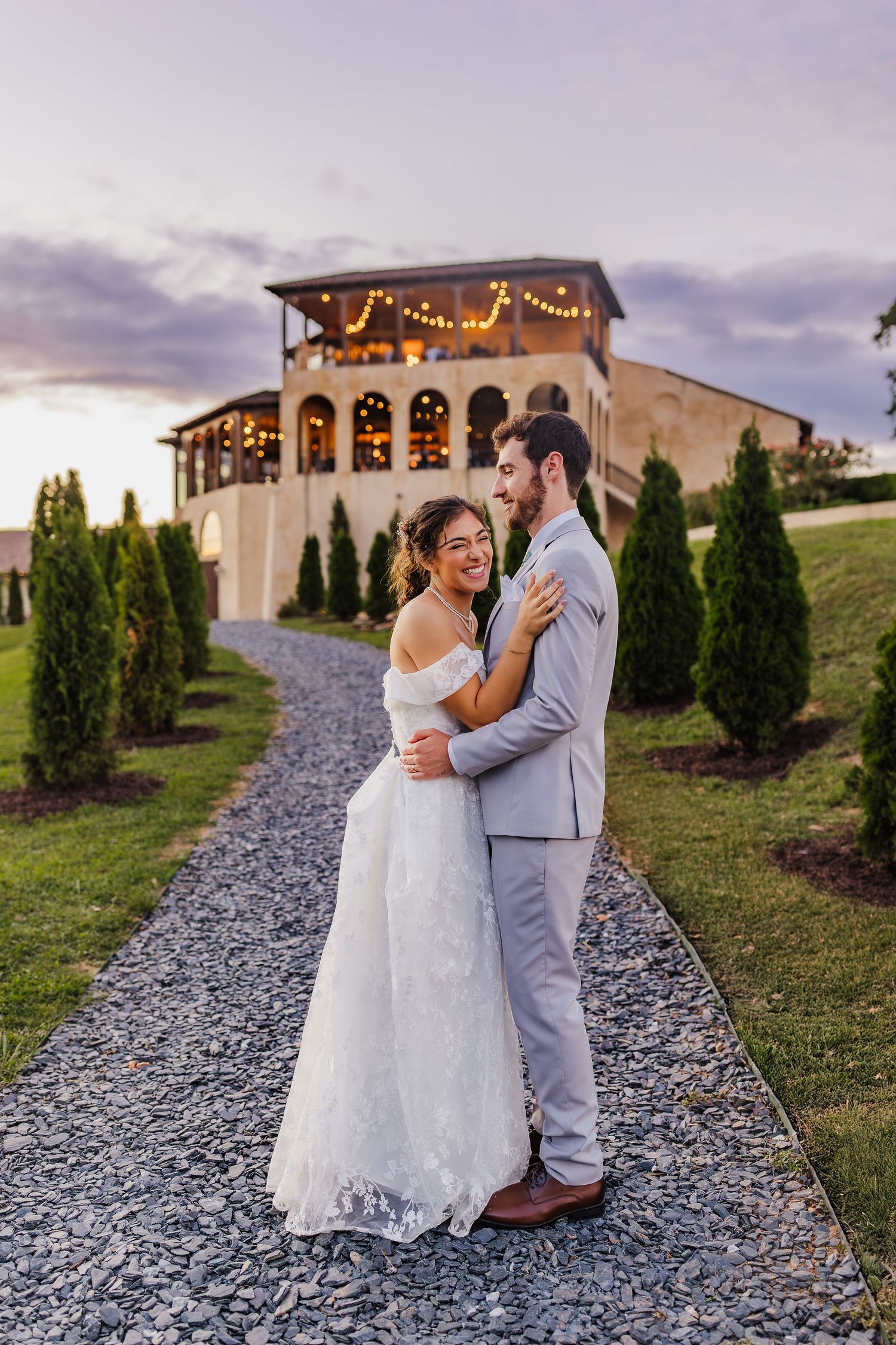 A bride and groom embrace on a gravel path at sunset, with a warmly lit wedding venue adorned with string lights behind them.
