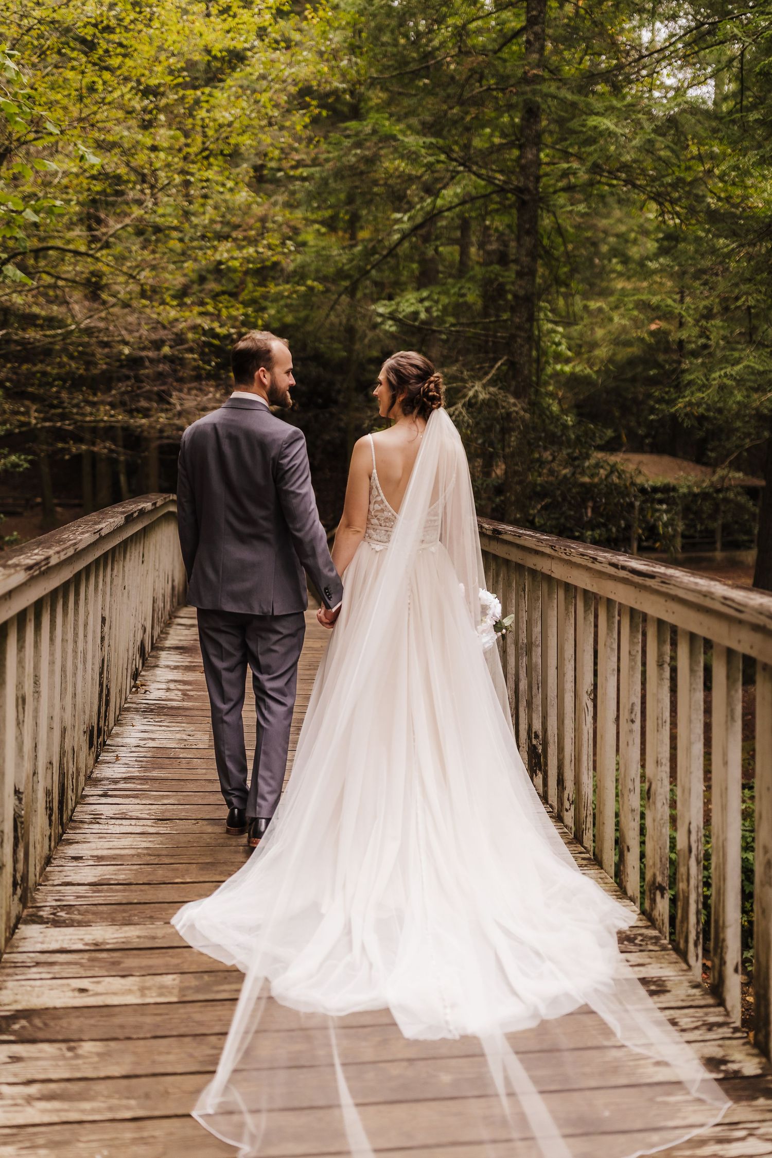 Bride and groom walking hand in hand across a wooden bridge surrounded by lush green trees on their wedding day.