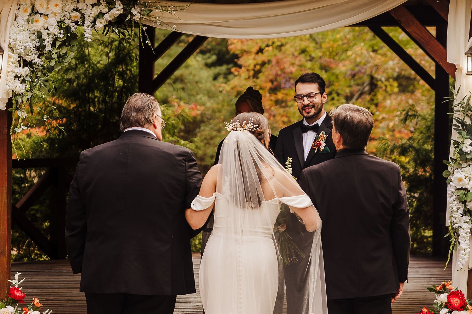 A wedding ceremony takes place outdoors under a white canopy with autumn foliage in the background.