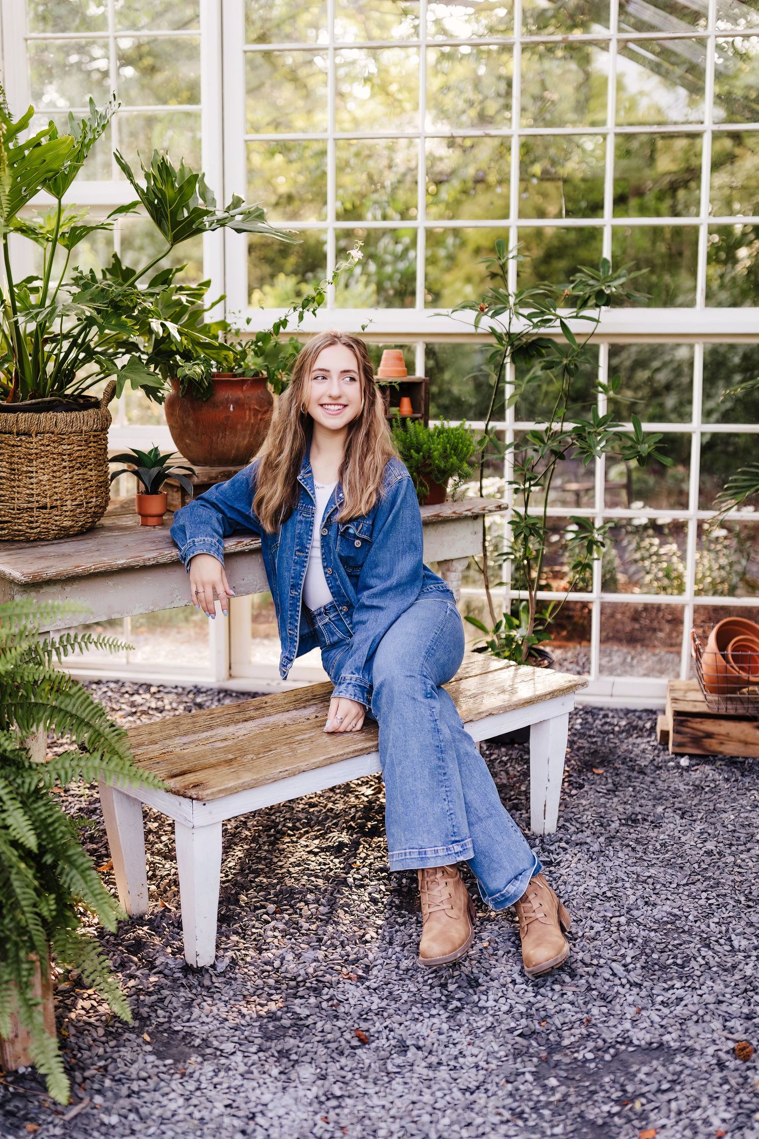 Young woman in denim outfit sitting on a wooden bench in a greenhouse surrounded by lush green plants.