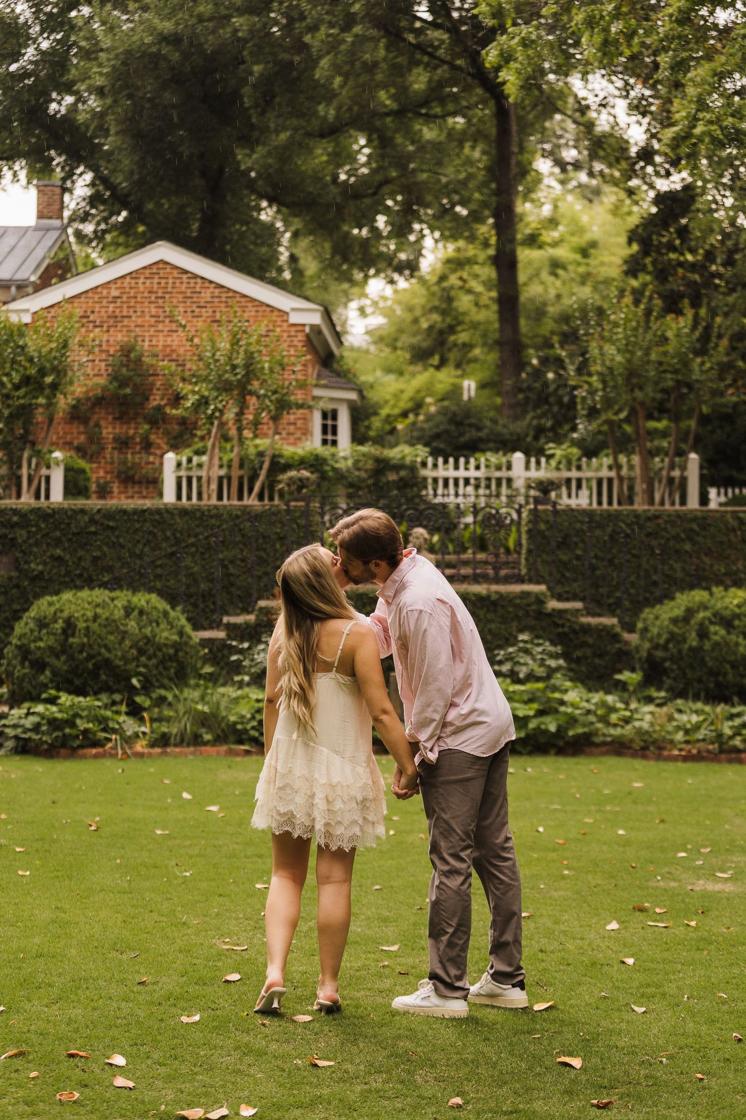 Couple holding hands and sharing a kiss in a lush garden with brick house and white fence backdrop.