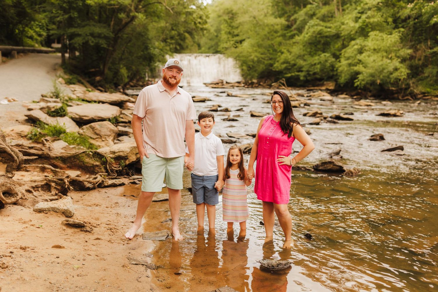 Family of four standing in shallow river water near rocky waterfall on sunny day.