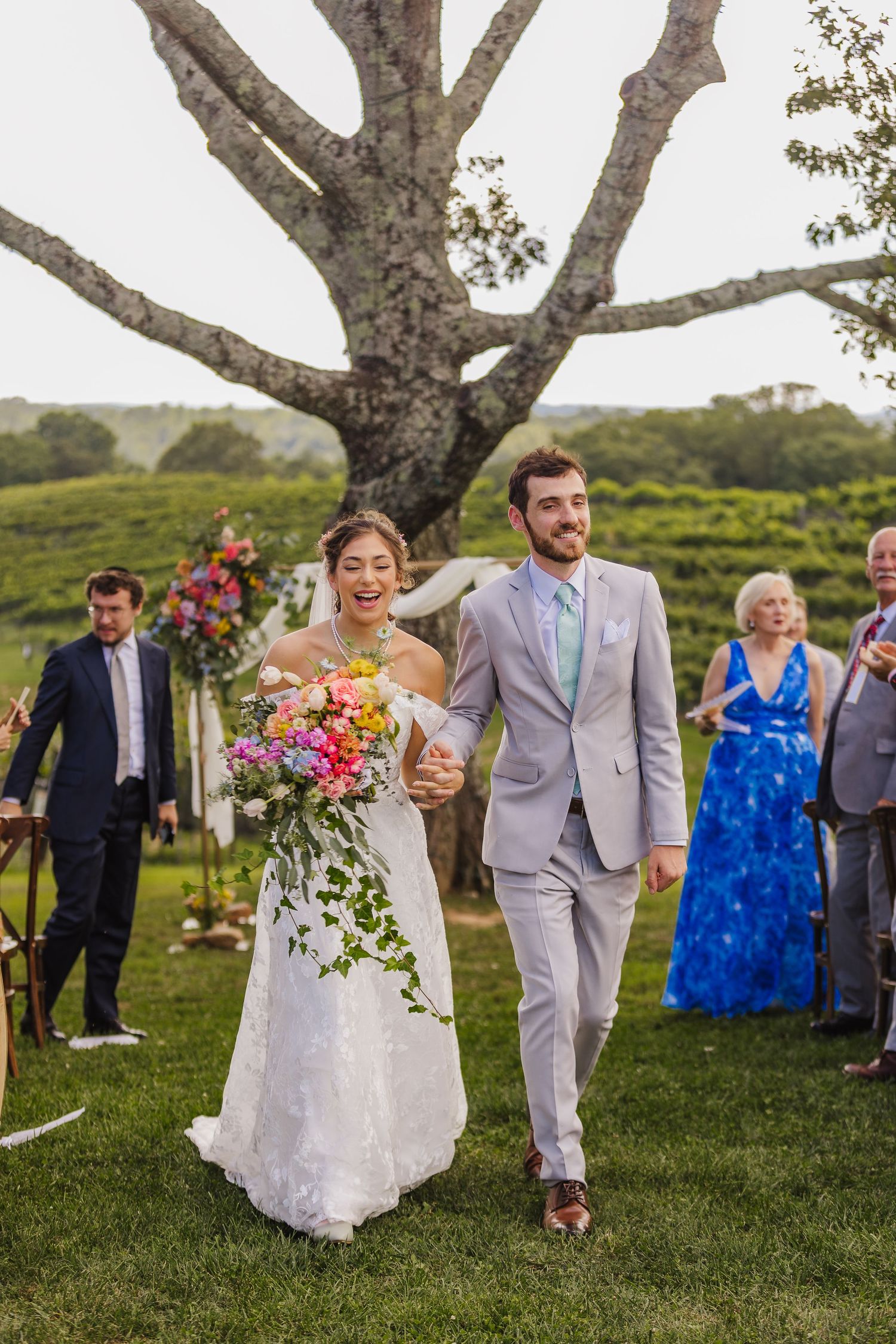 Bride and groom walk down the aisle laughing, she holds colorful bouquet, he wears gray suit, outdoor vineyard wedding.