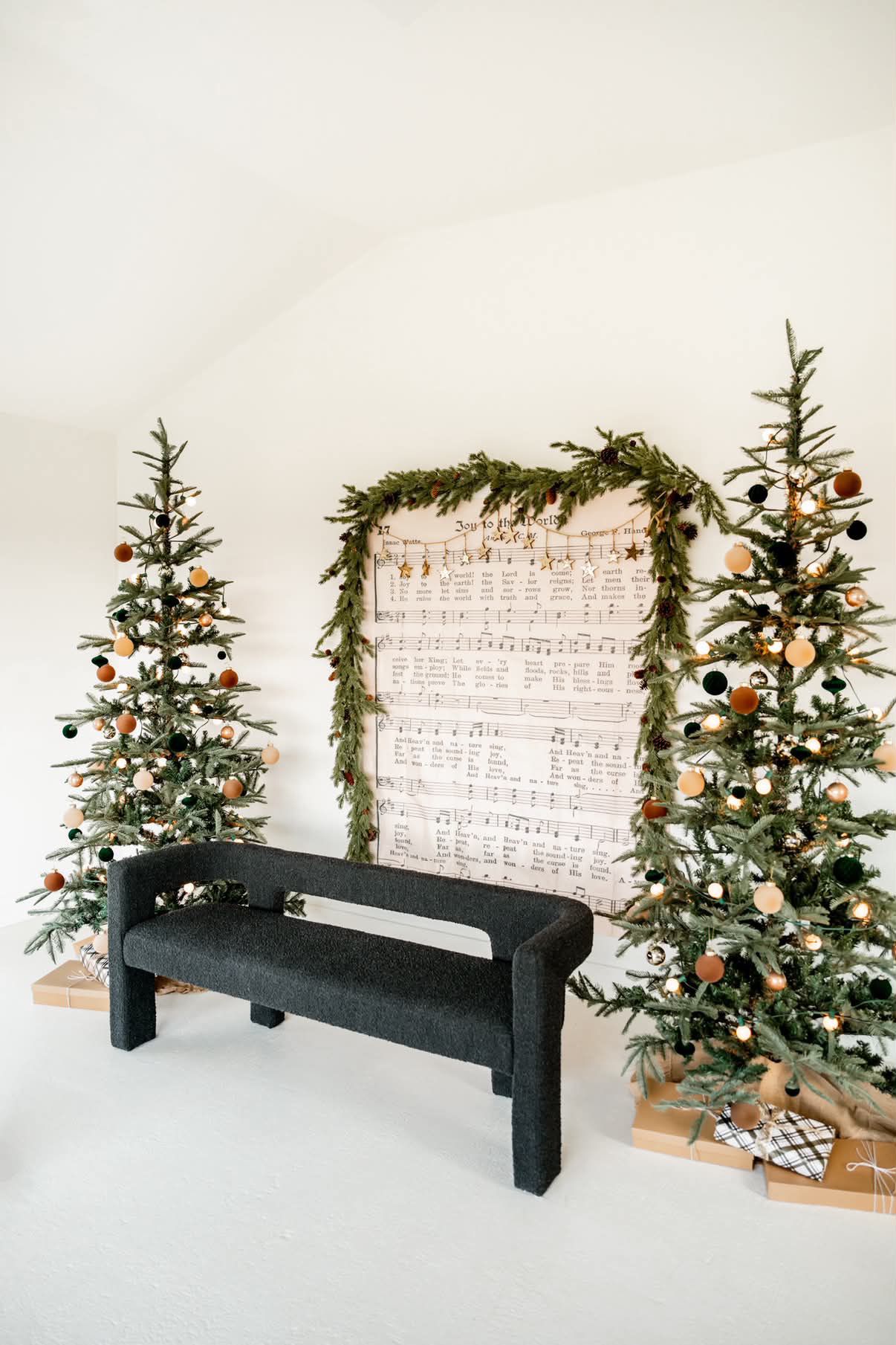 Two slim Christmas trees with natural ornaments flank a sheet music backdrop with garland, above a black bench.