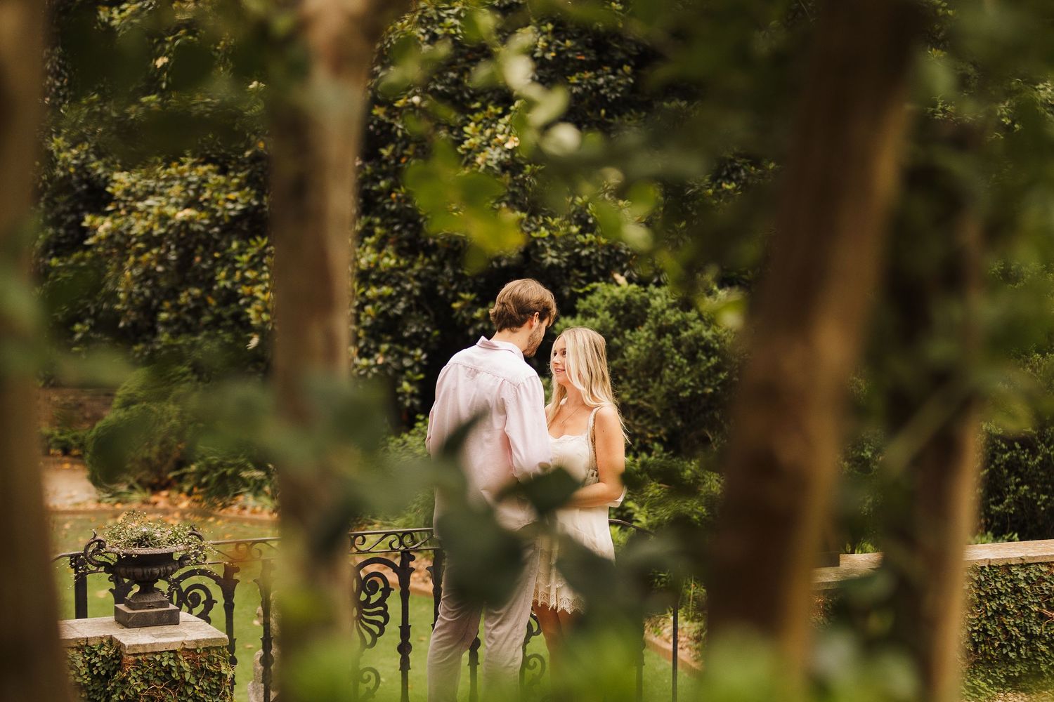 Couple standing together in a lush garden surrounded by green trees and manicured hedges during an outdoor engagement session.