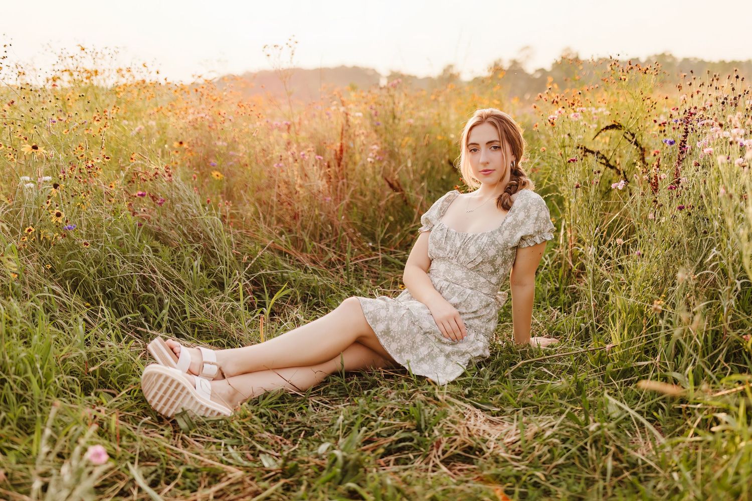 Young woman in floral dress sitting in a wildflower meadow during golden hour sunset.