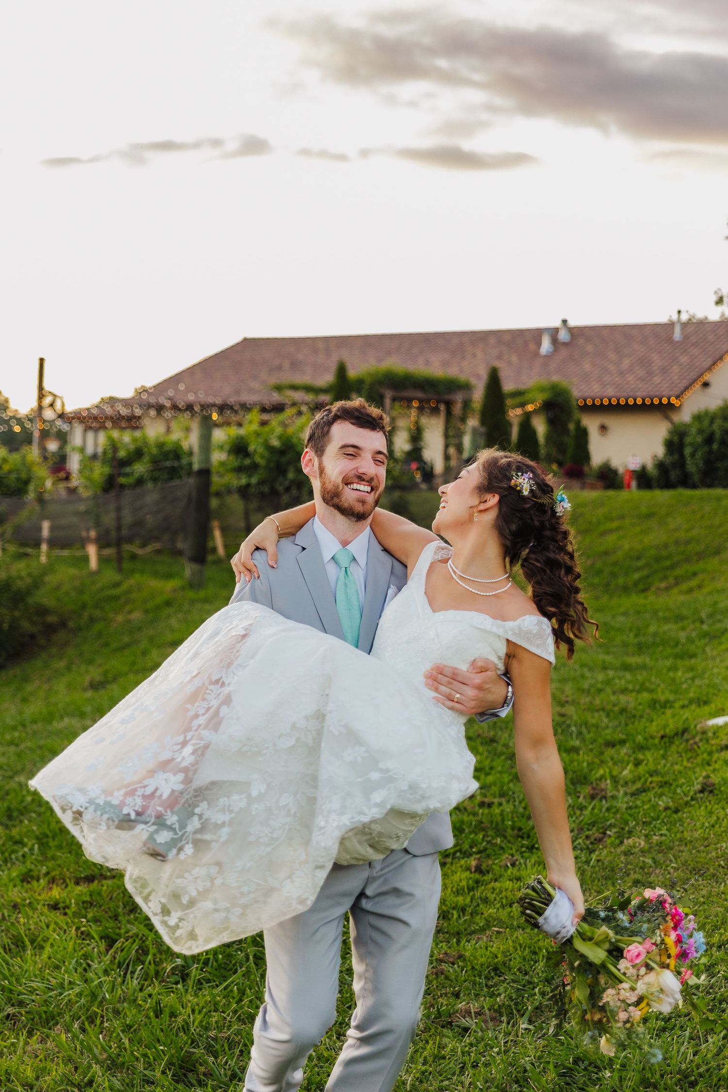 Smiling groom carries bride in white wedding dress on lush green lawn with vineyard and rustic building backdrop.