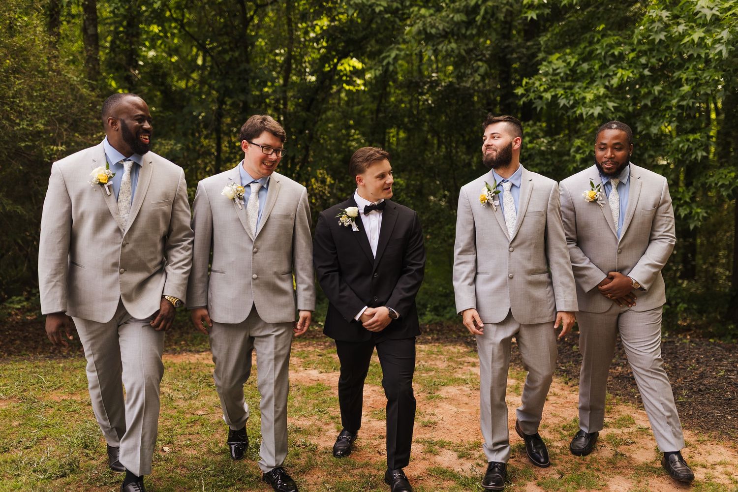 Groom in black tuxedo stands with four groomsmen in matching gray suits and boutonnieres outdoors.