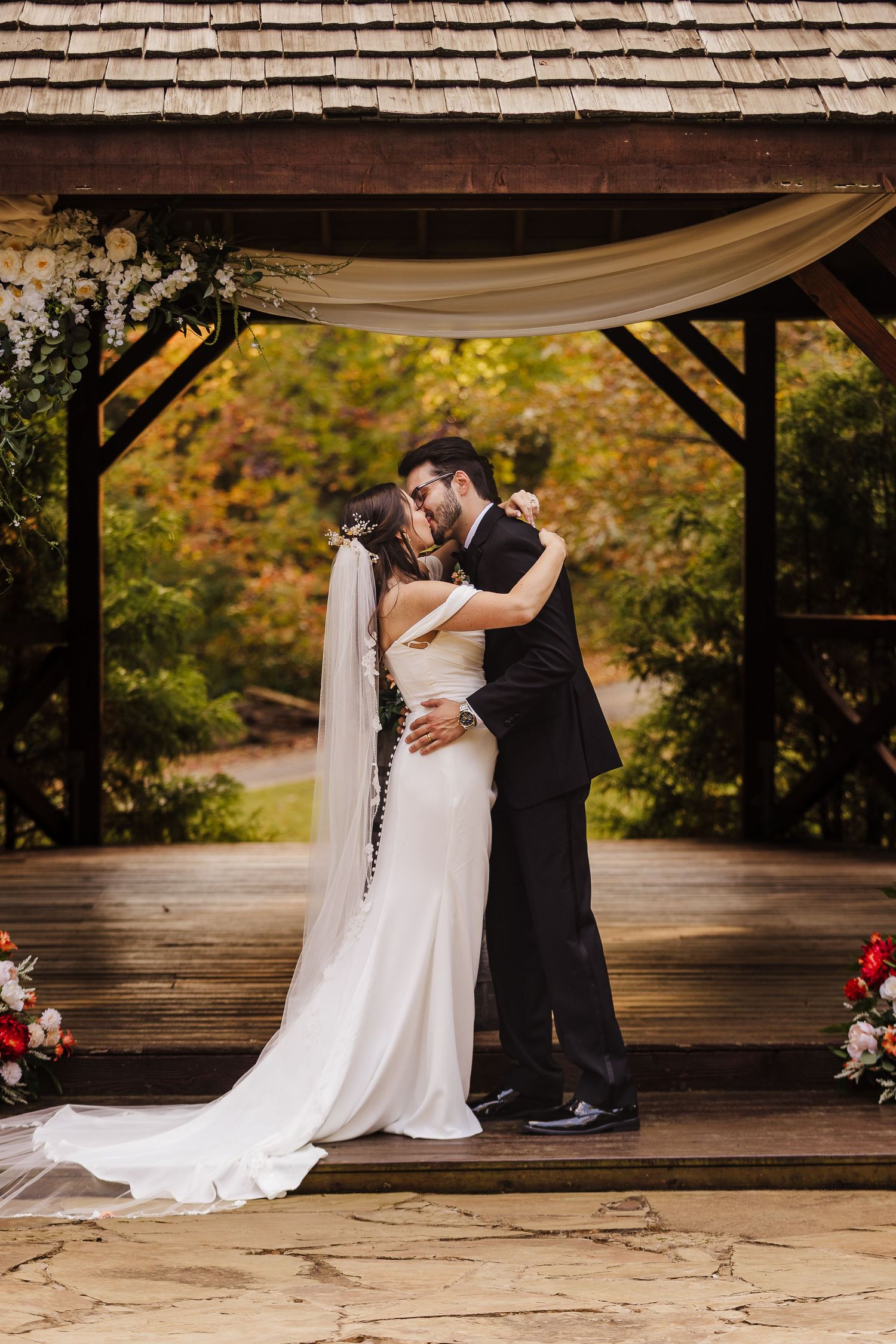 A romantic wedding ceremony takes place under a rustic wooden pavilion with autumn foliage in the background.