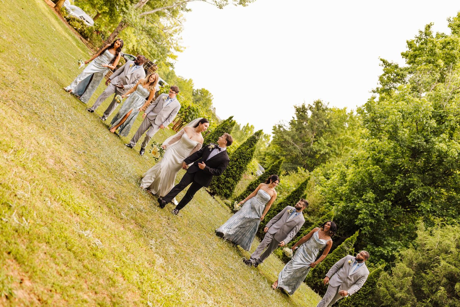 Wedding party walking on a grassy hillside outdoors, bridesmaids in silver gowns and groomsmen in gray suits.