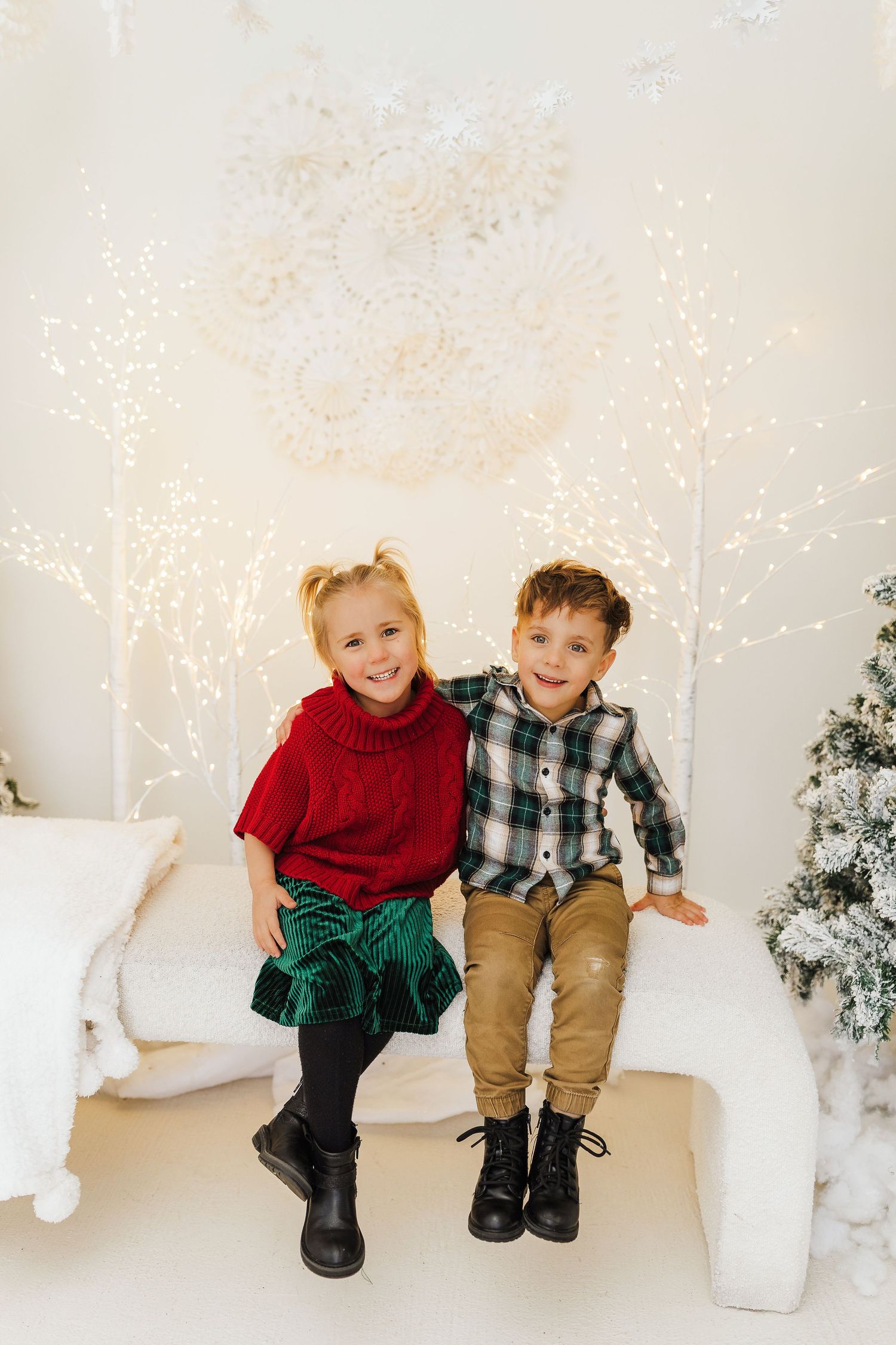 Two young children in holiday outfits sit together near a Christmas tree and festive white lights.